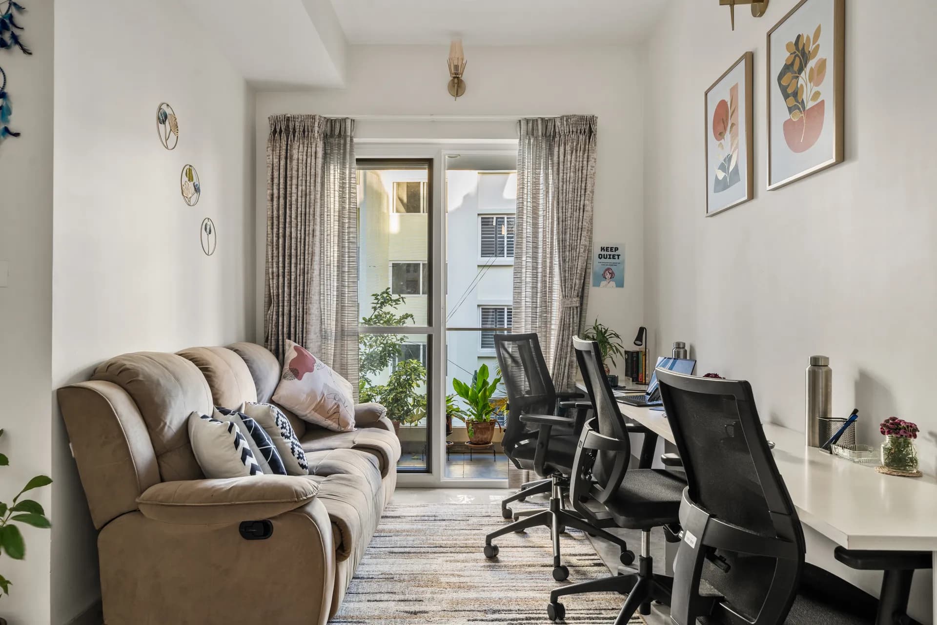 Bright workspace with a desk and two black task chairs, a beige recliner sofa, and a glass door opening onto a balcony with potted plants.
