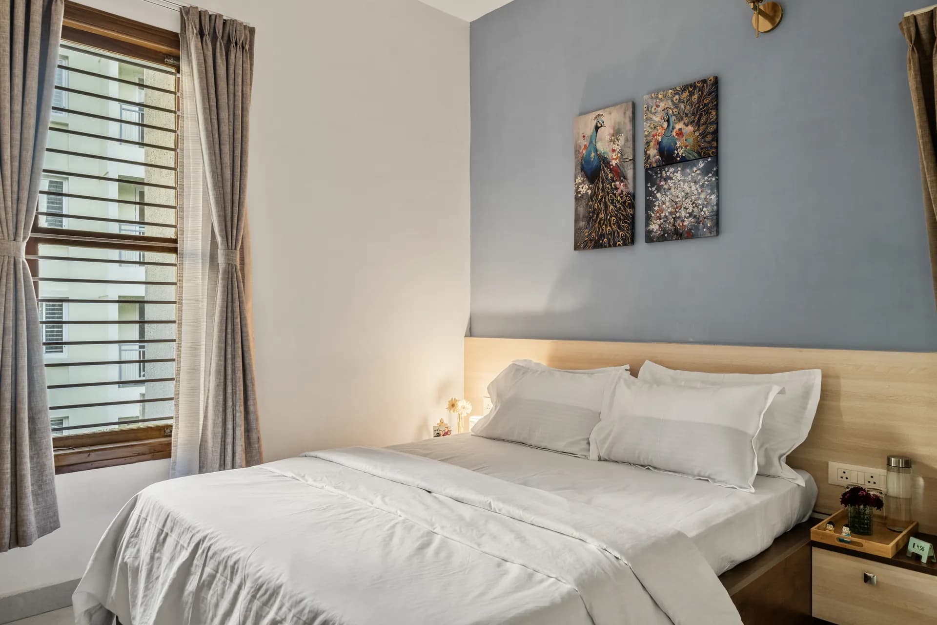 Bedroom with a made-up queen bed, blue accent wall with peacock artwork, and a window with grey curtains.