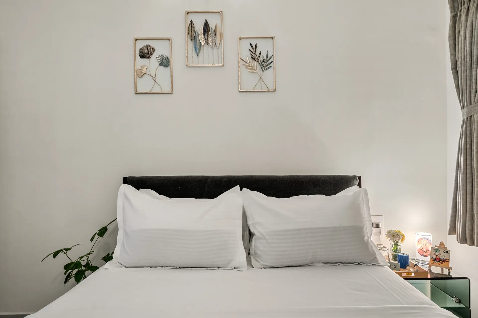 Bedroom with a made bed of white linen, dark upholstered headboard, framed leaf wall art and a small bedside lamp.