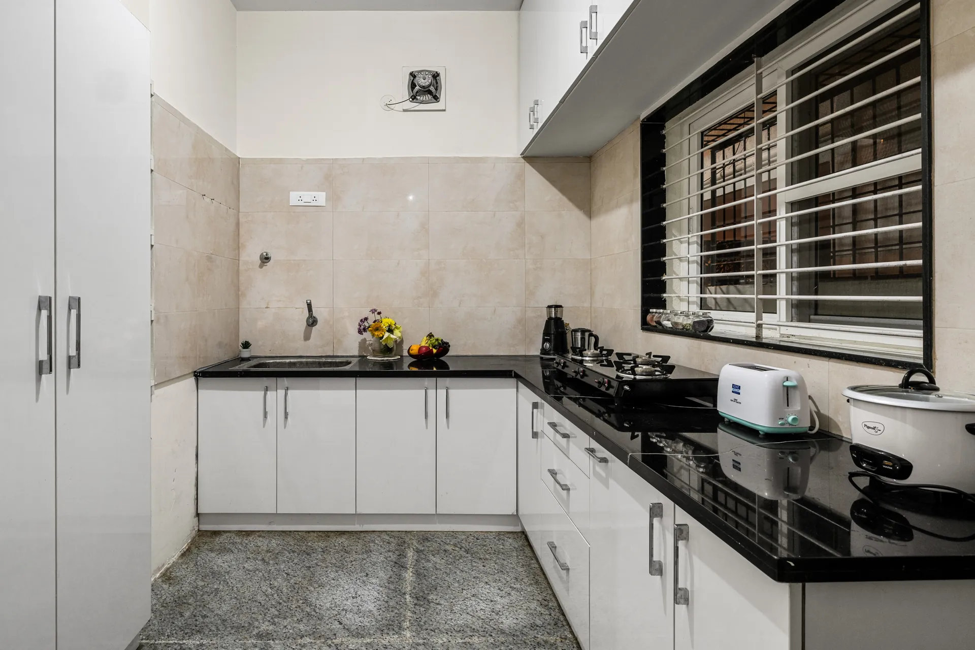 Galley-style kitchen with black granite counters, a two-burner gas hob, toaster and slow cooker, lit by a barred window.
