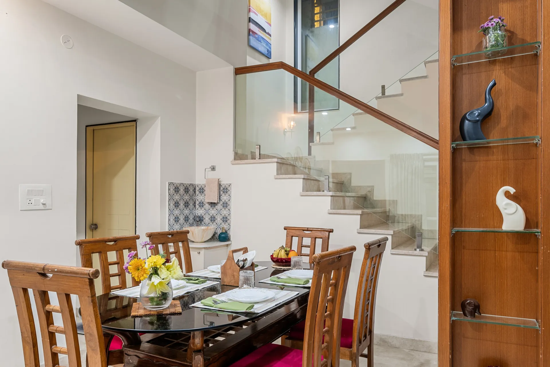 Dining area with a glass-top table set for four, flowers in a vase, and a glass-railed staircase behind.