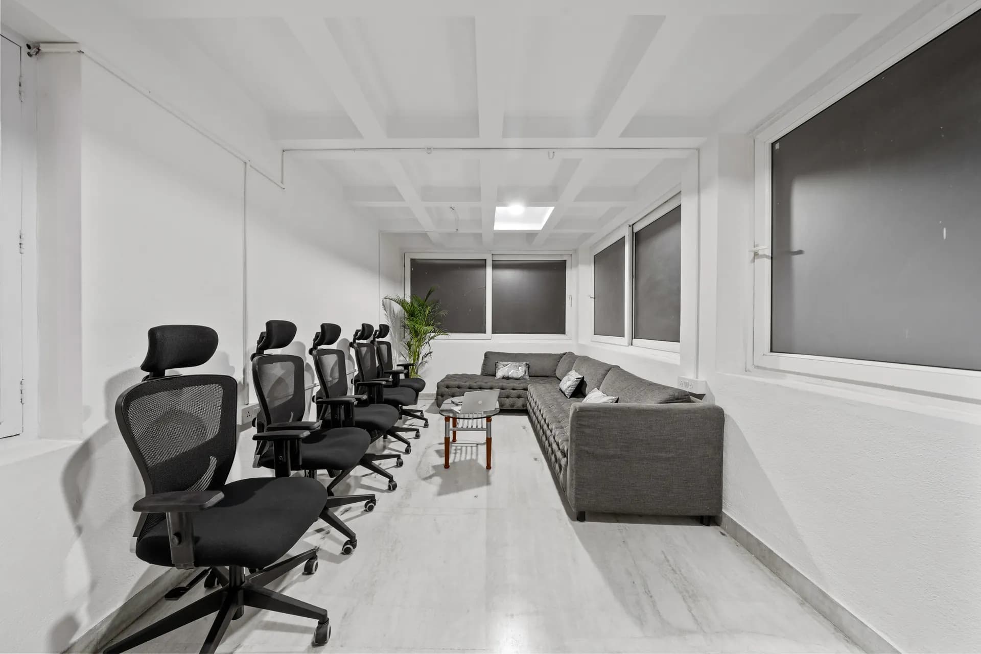 A bright white lounge with a row of black mesh office chairs, a grey sectional sofa and a potted palm.