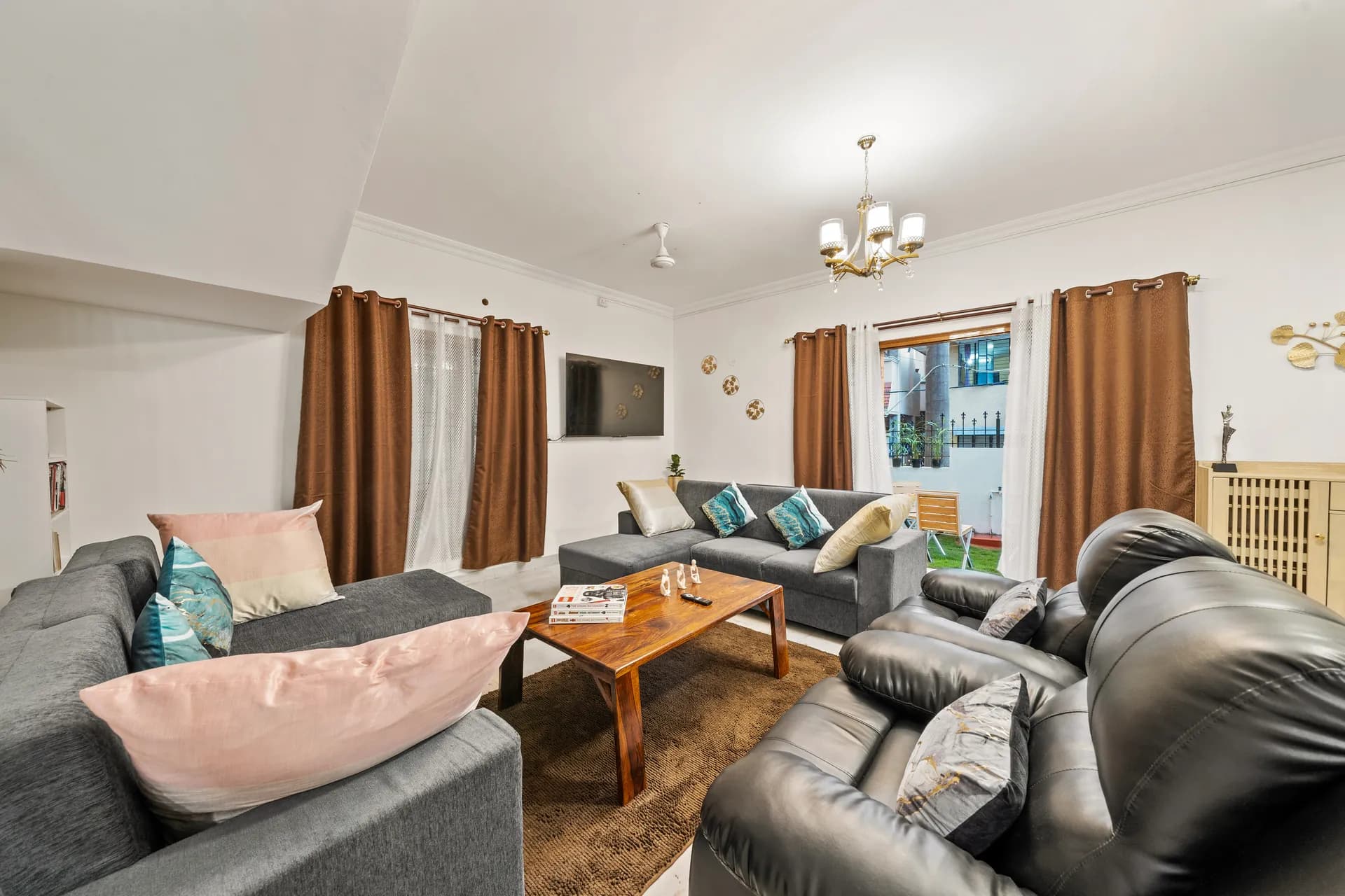 Living room with grey sectional, leather recliners, wooden coffee table, chandelier and TV; garden visible through the window.