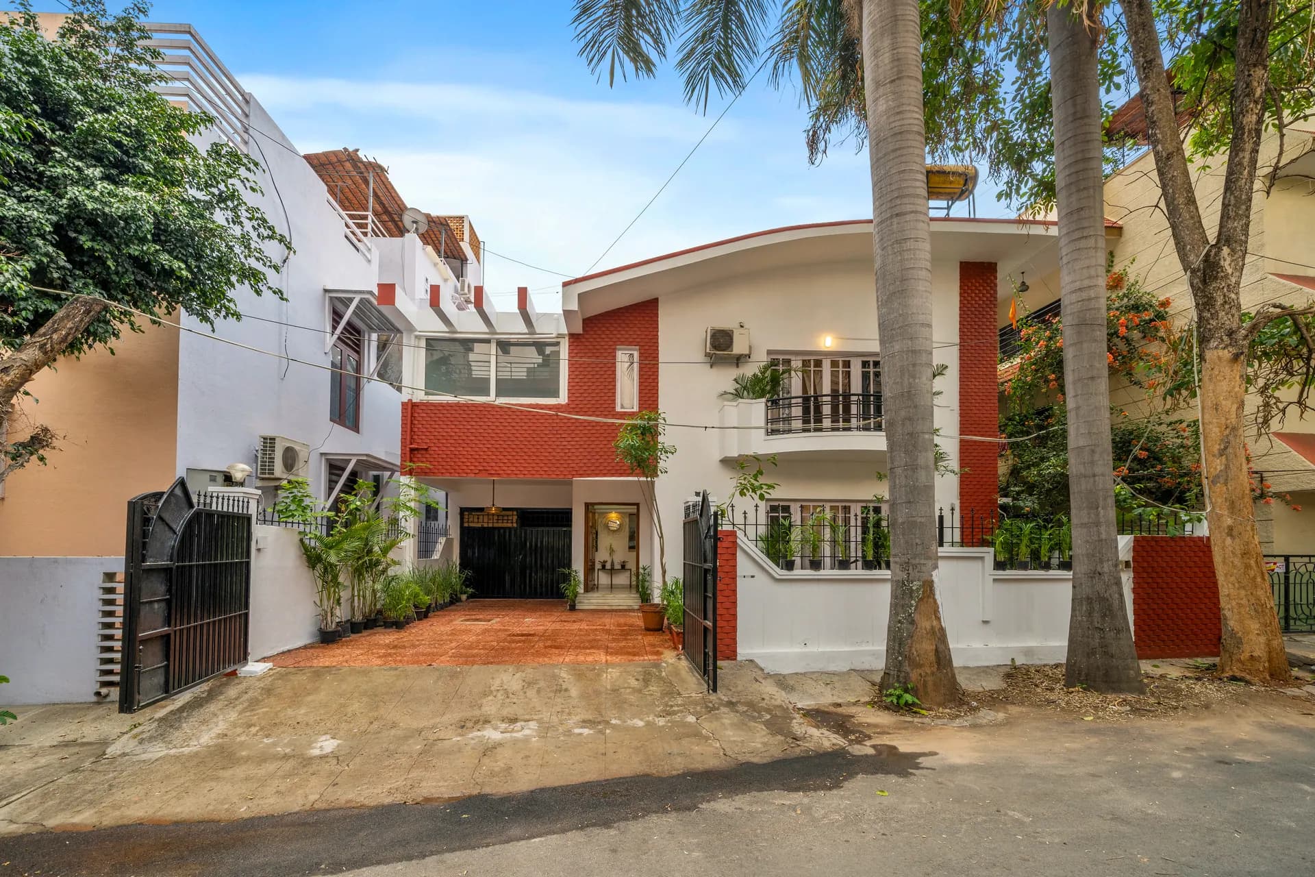 Front of a two-storey red-and-white villa with a covered parking area, potted plants and an iron gate.