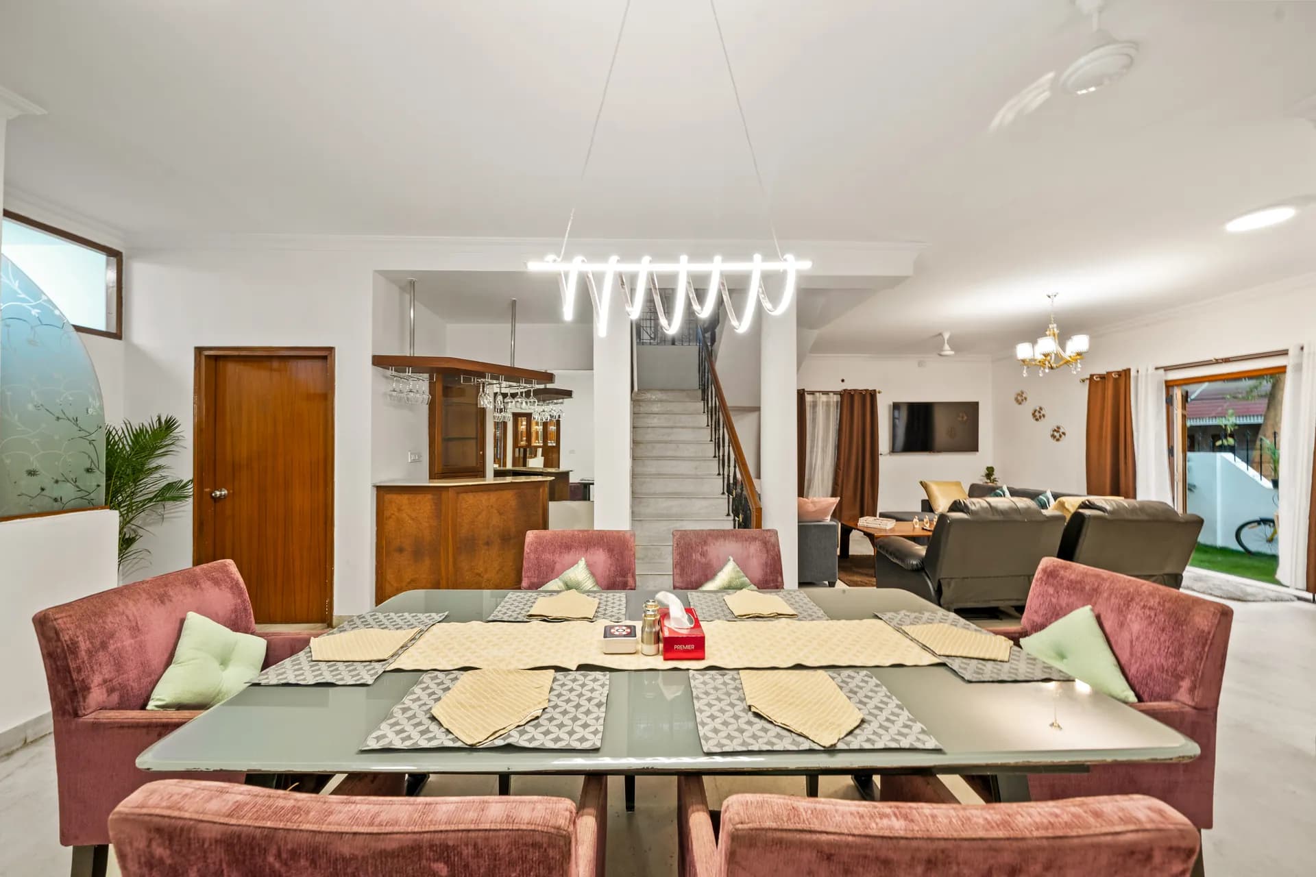 Open-plan dining area with a long glass table, six pink upholstered chairs, and a sofa lounge visible beyond.