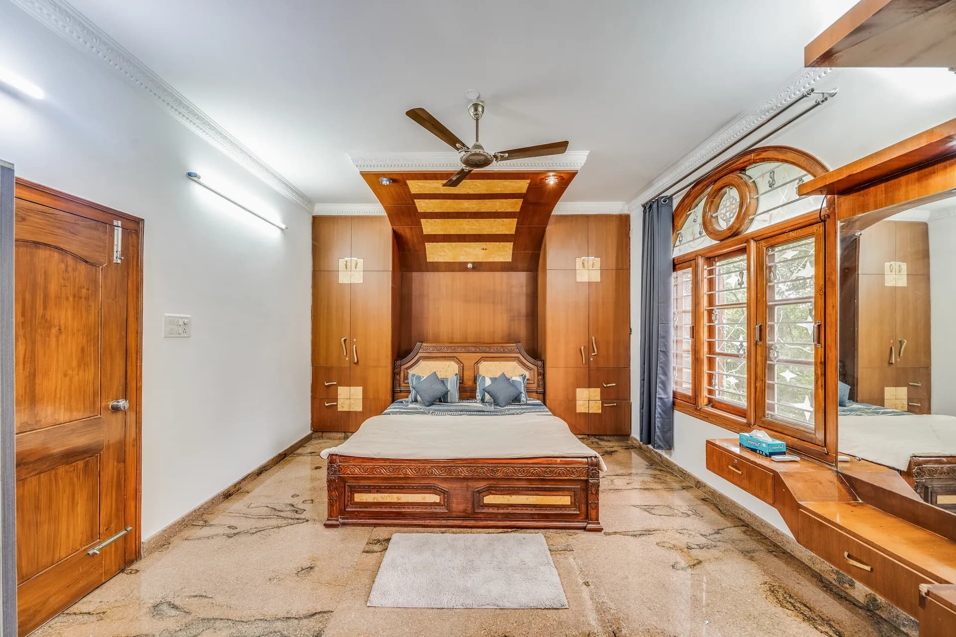 Bedroom with a wood-framed double bed, wall-to-wall wardrobes, a ceiling fan and an arched window with a dressing unit.