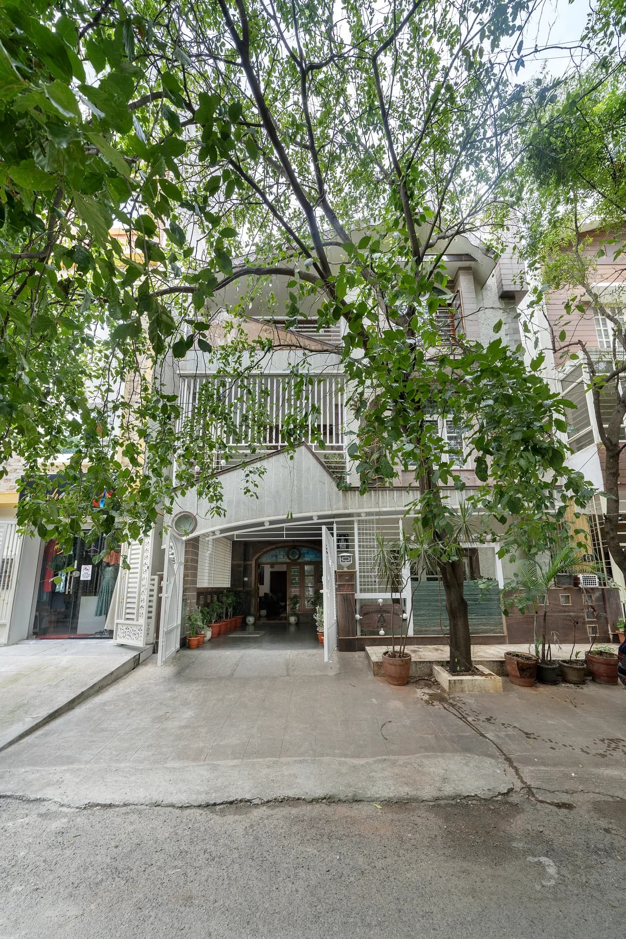 Front of a three-storey villa with a covered entrance, potted plants and a large tree shading the driveway.