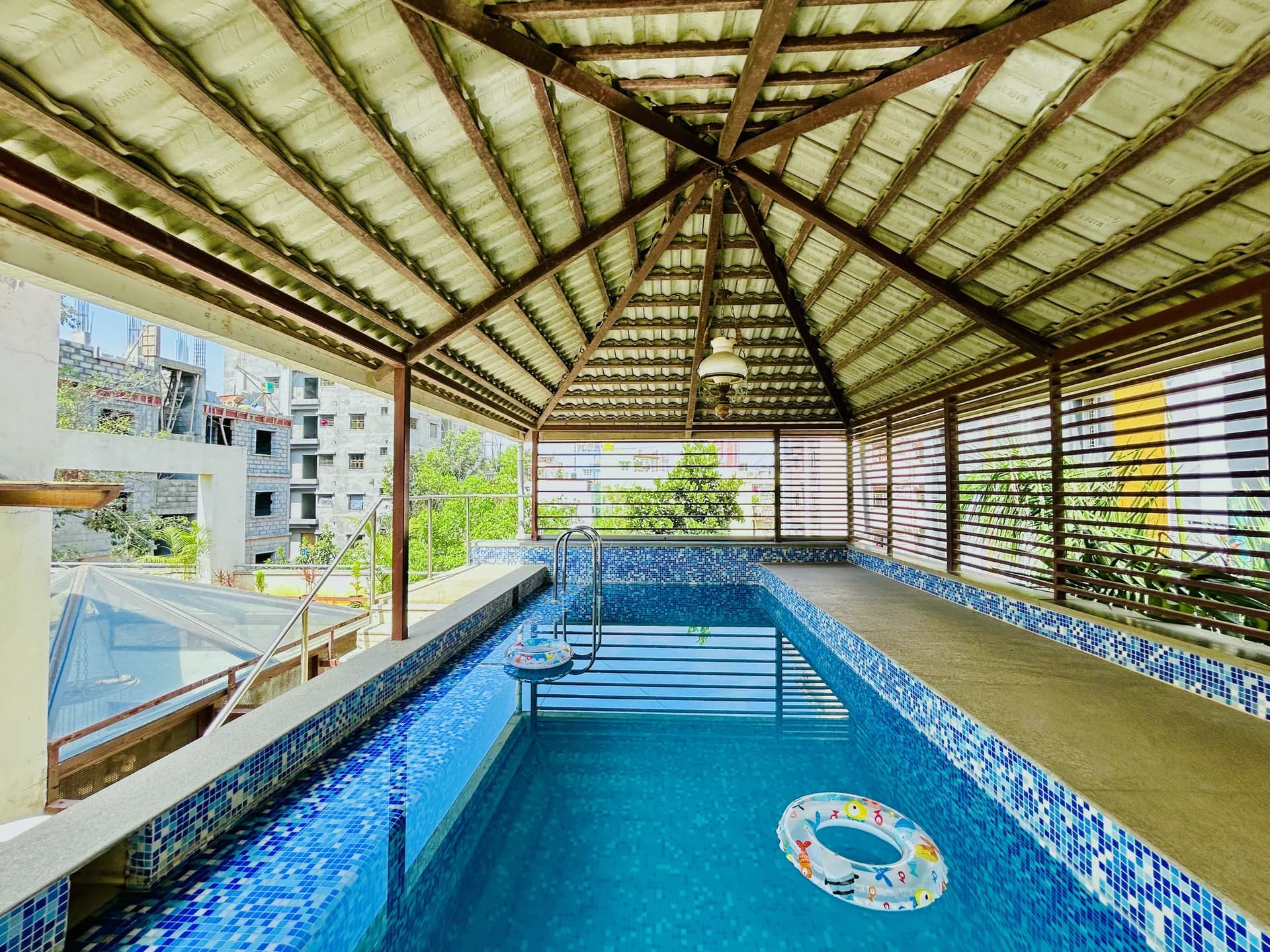 Covered indoor swimming pool with blue mosaic tiles, a wooden slatted roof, and a pool ring floating in the water.