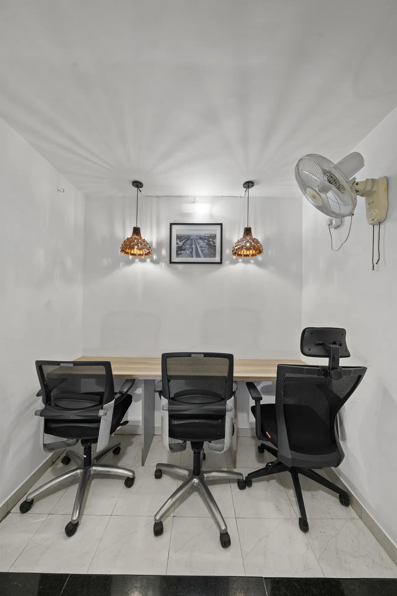 Small workspace room with a wooden desk, three black mesh office chairs, two pendant lamps and a wall fan.