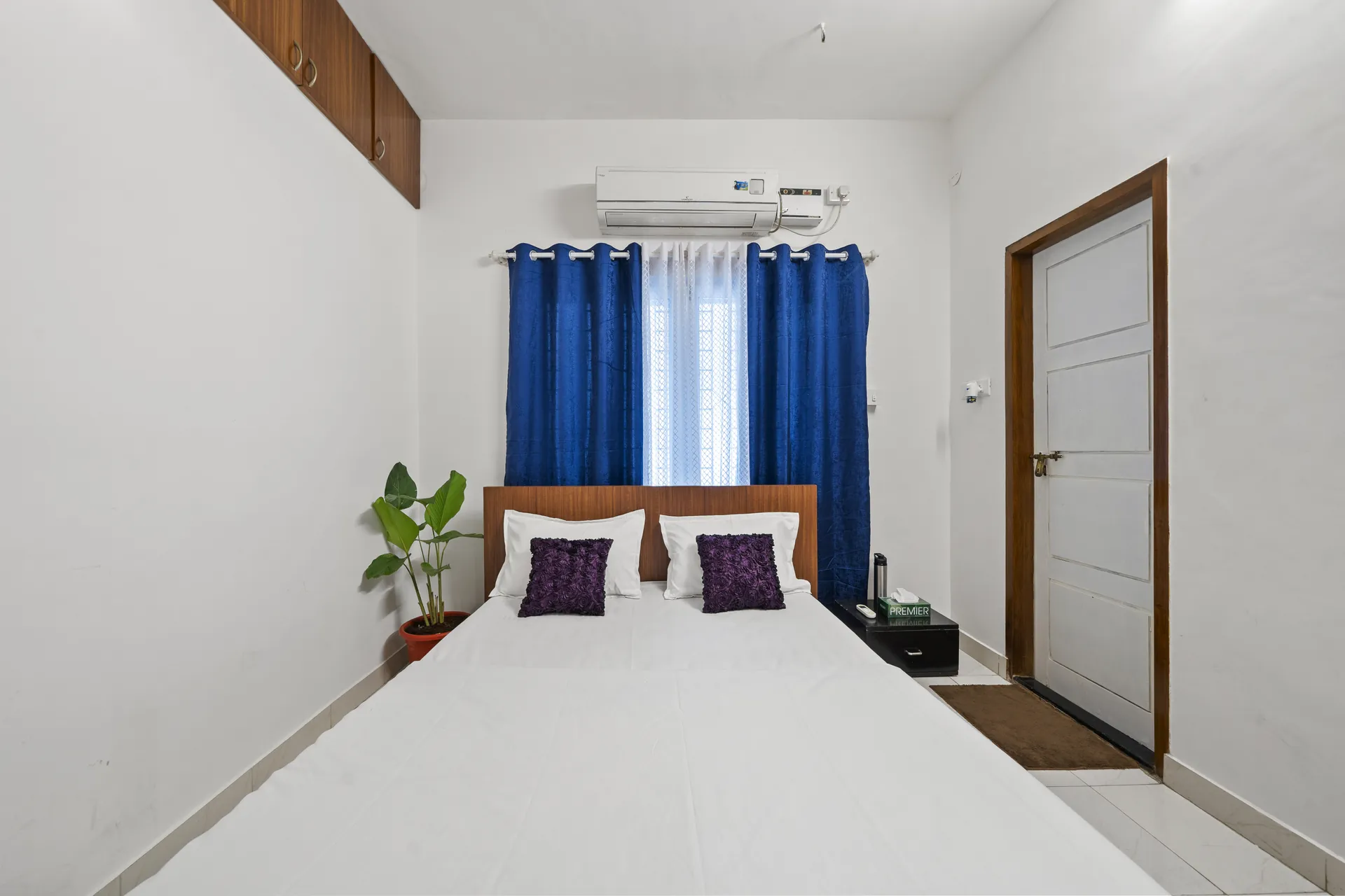 Bedroom with a made double bed, blue curtains, wall-mounted air conditioner and a potted plant by the bed.