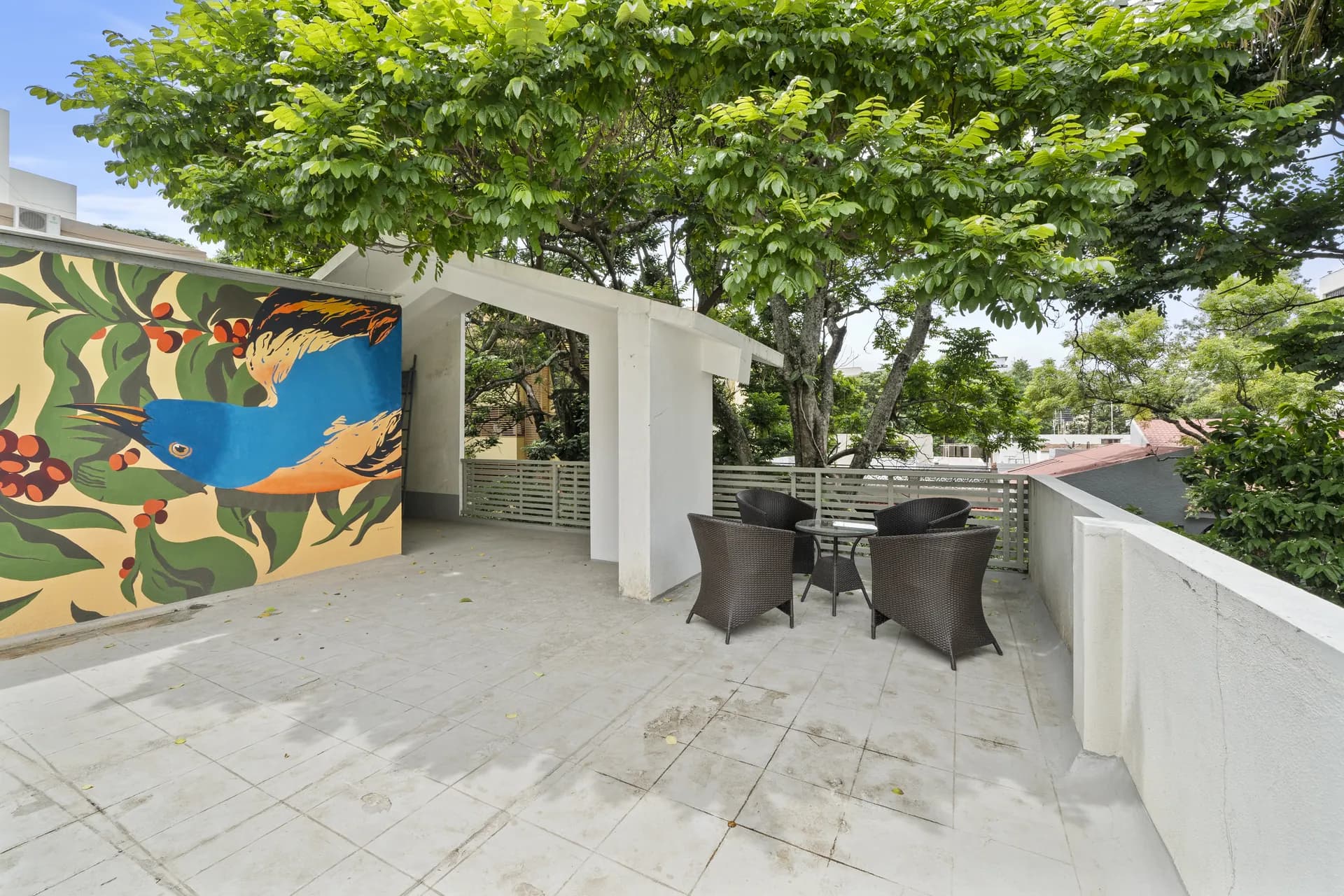 Covered terrace with a painted blue bird mural, a round table and four wicker chairs under a large tree.