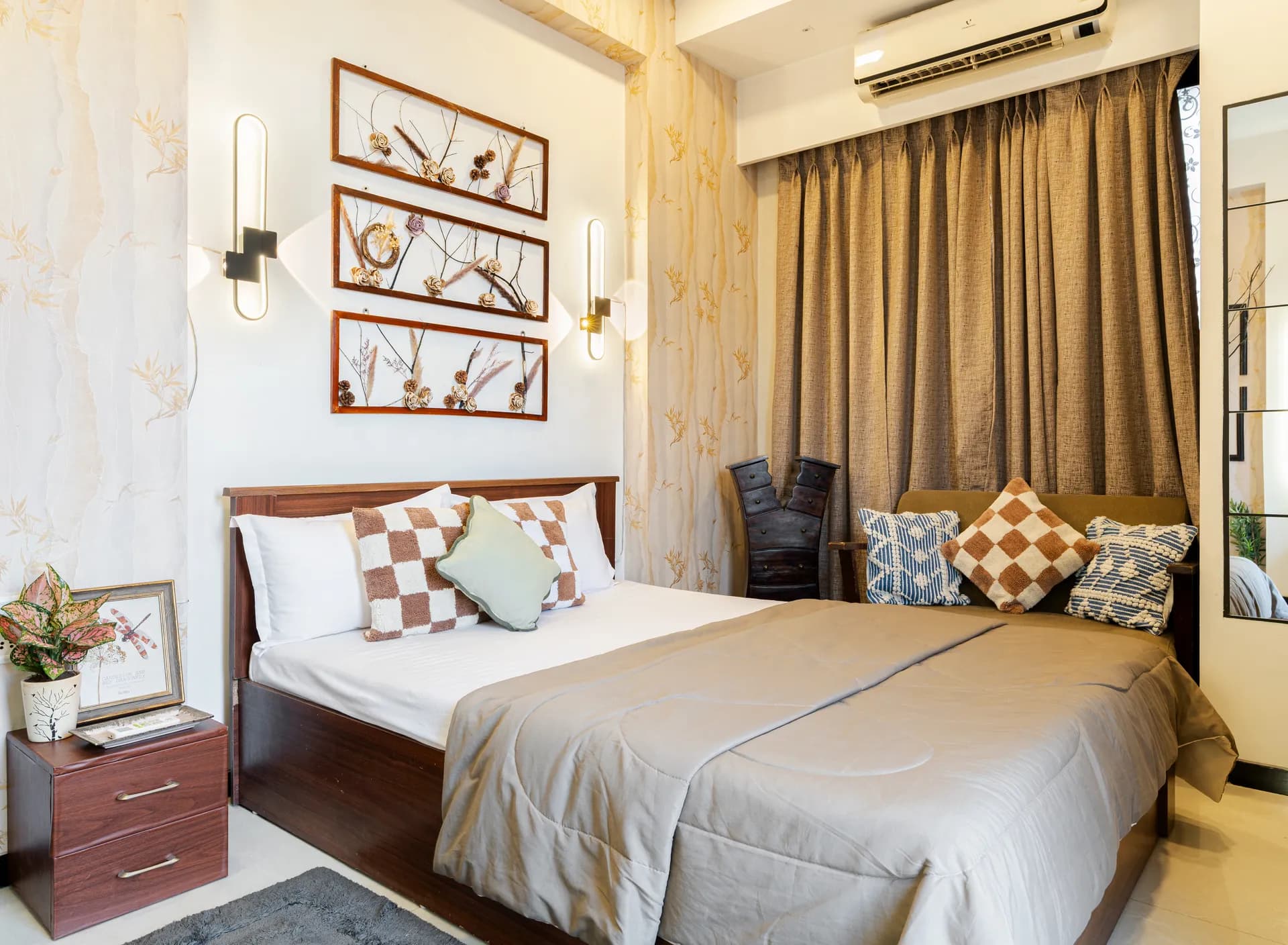 Bedroom with a double bed, layered cushions, wall art panels and a wall-mounted air conditioner above the curtains.