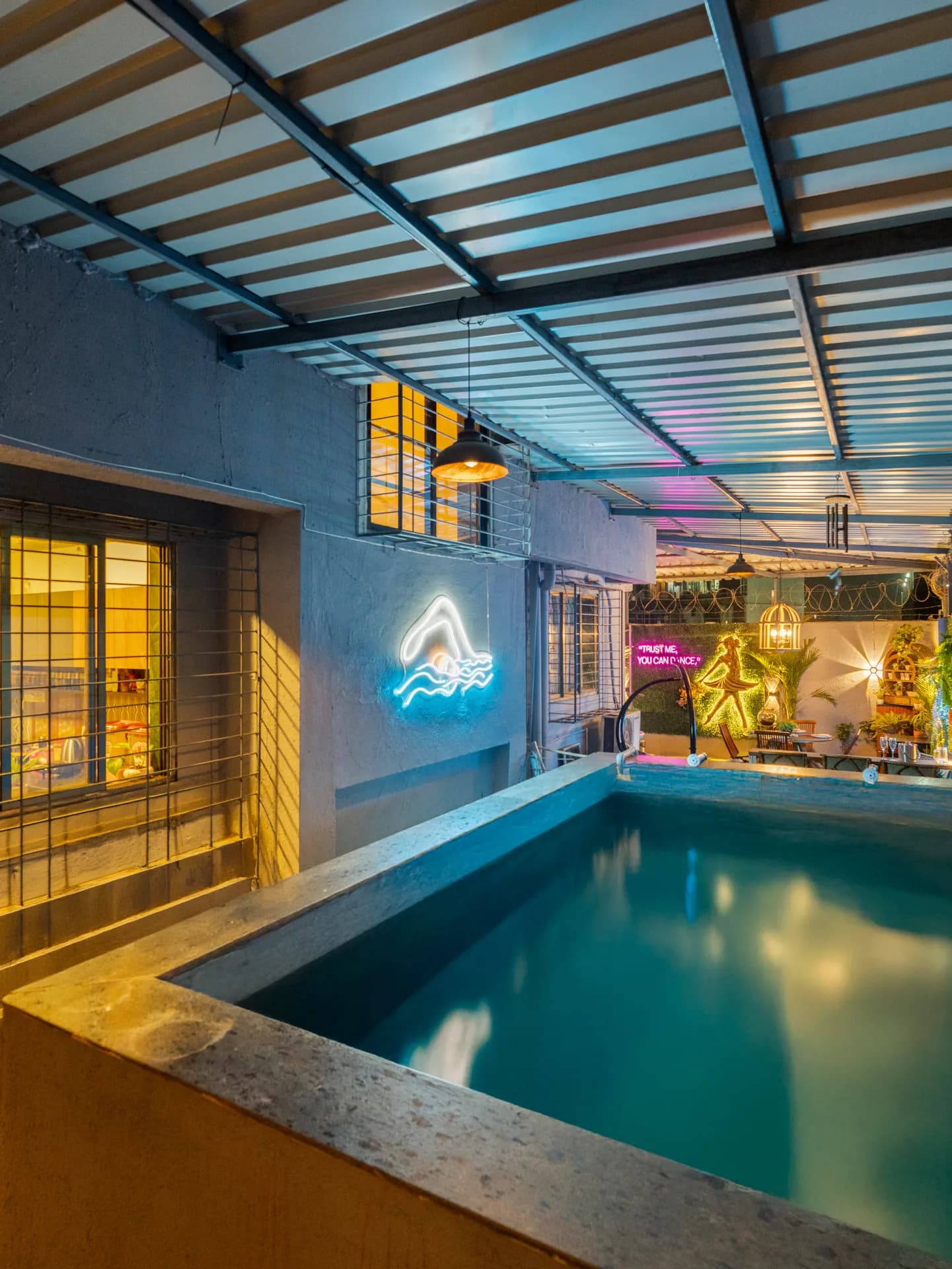 Covered outdoor pool under a corrugated roof, with neon signs and a lit garden bar area beyond.