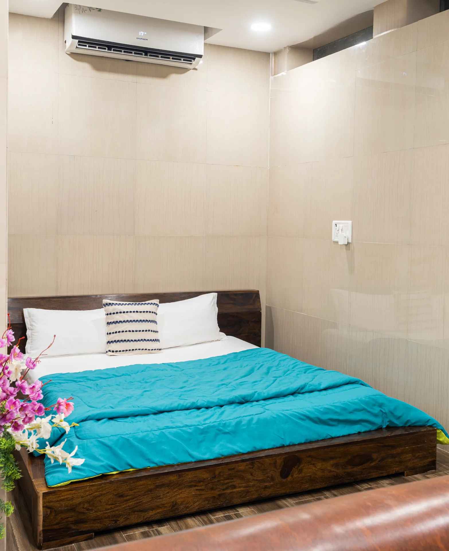 Bedroom with a wooden platform bed, teal duvet, wall-mounted air conditioner and pink flowers in the foreground.