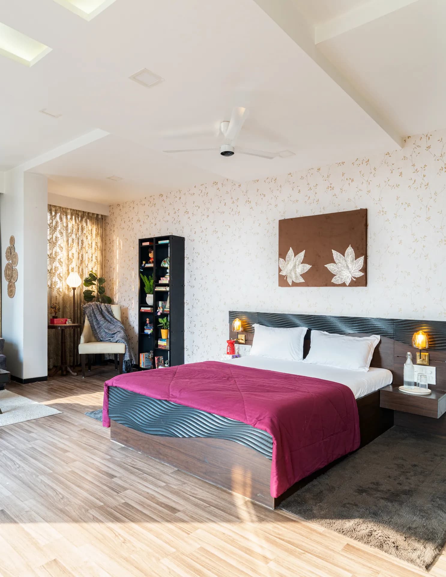 Bedroom with a made-up king bed under a magenta throw, a reading chair by the curtains, and a tall bookshelf.