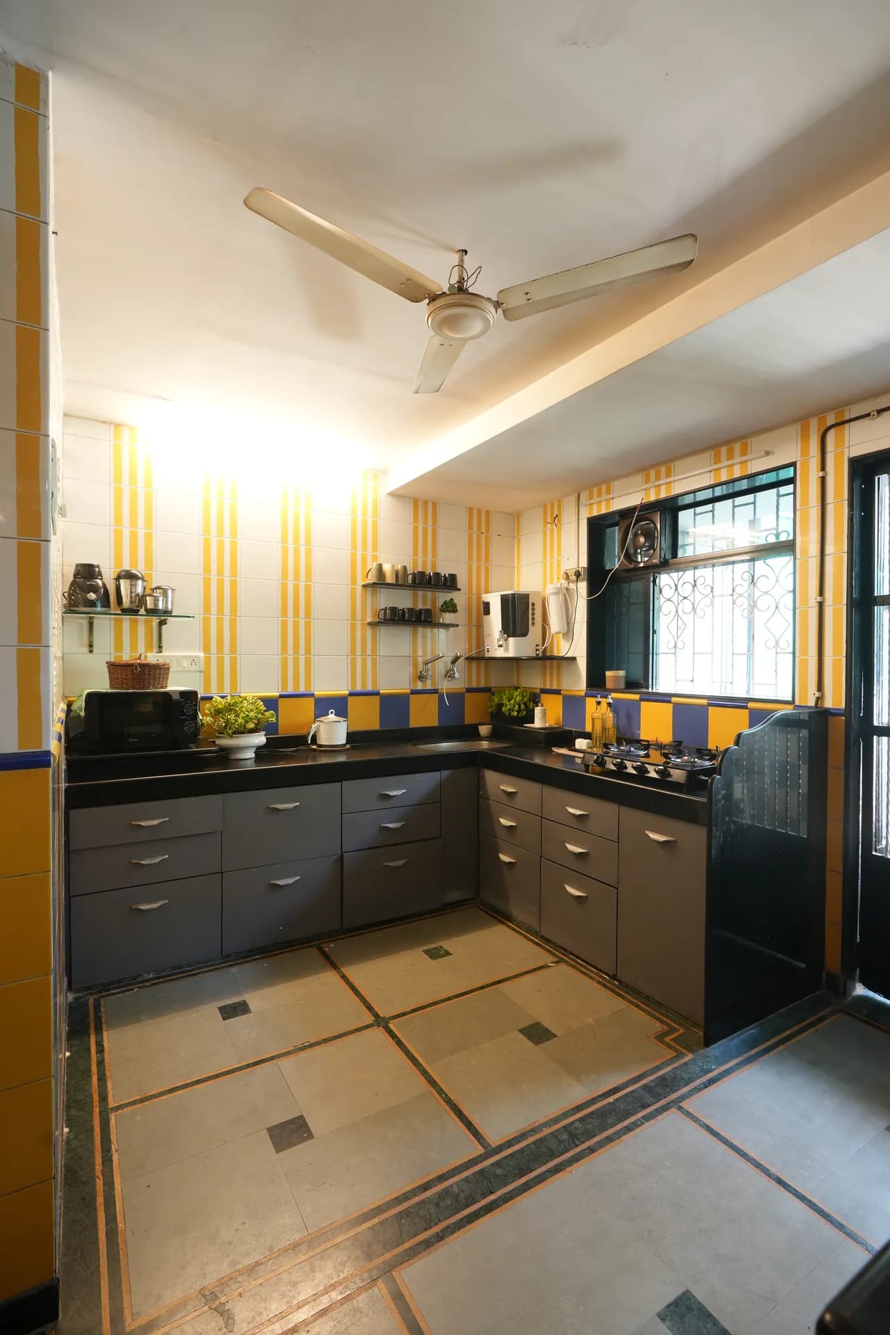 Galley-style kitchen with grey cabinets, black countertops, yellow-and-blue tiled walls, and a ceiling fan.