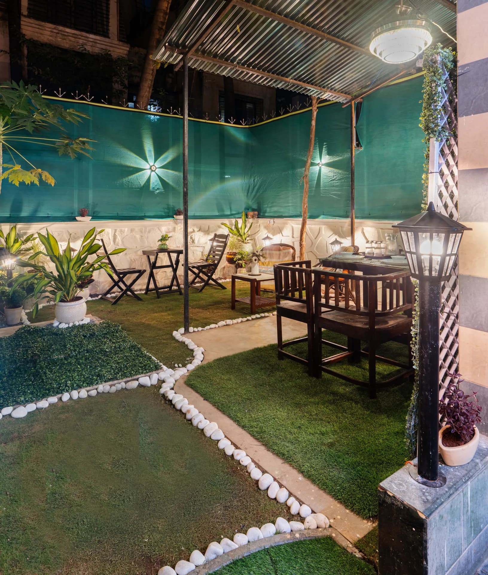 Covered garden terrace at night with dark wooden dining chairs, folding chairs, potted plants and white pebble-edged artificial lawn.