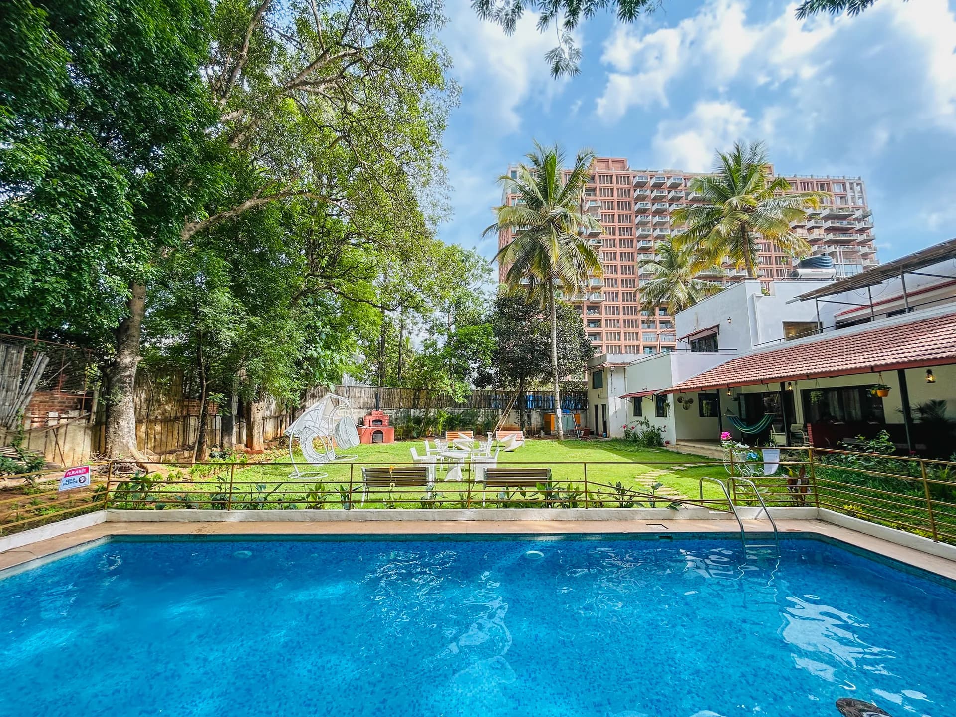 Blue swimming pool in a leafy garden, with a villa terrace and lawn behind it and a tall apartment block in the distance.