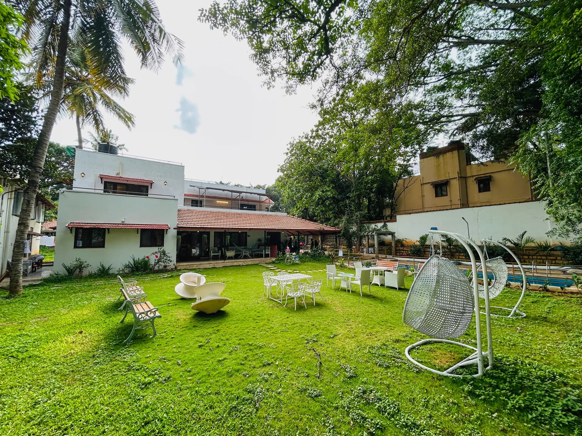 Lawn garden with white bench seating, round white chairs, egg-shaped hanging swing chairs, and villa behind.