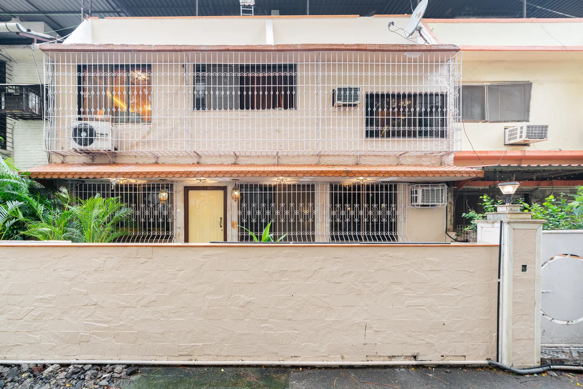 Front facade of a two-storey house behind a tall cream wall, with barred windows, a yellow door and wall-mounted air conditioners.