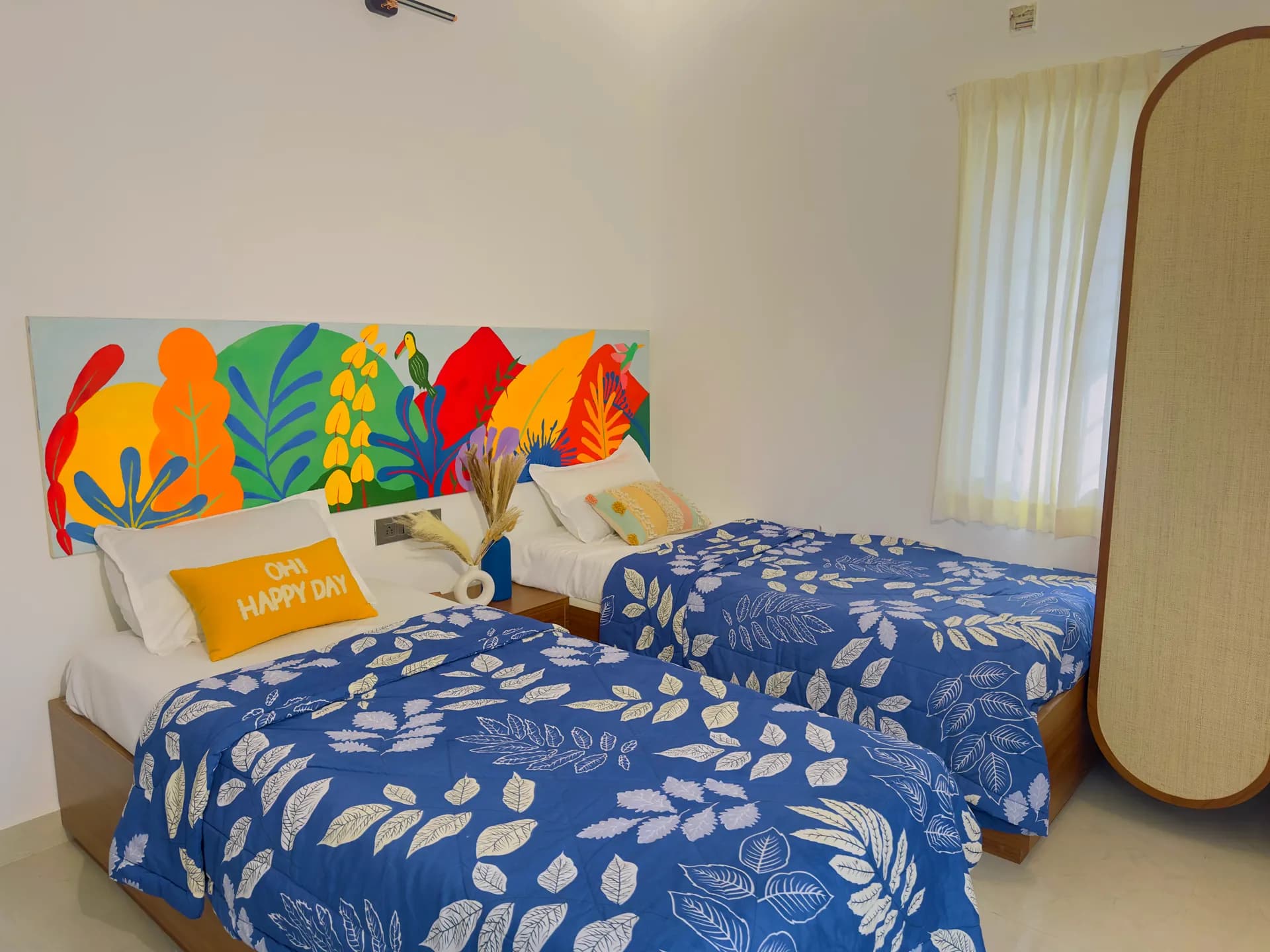 Bedroom with two single beds under a colourful painted headboard, blue leaf-print covers, and a tall wardrobe by the window.