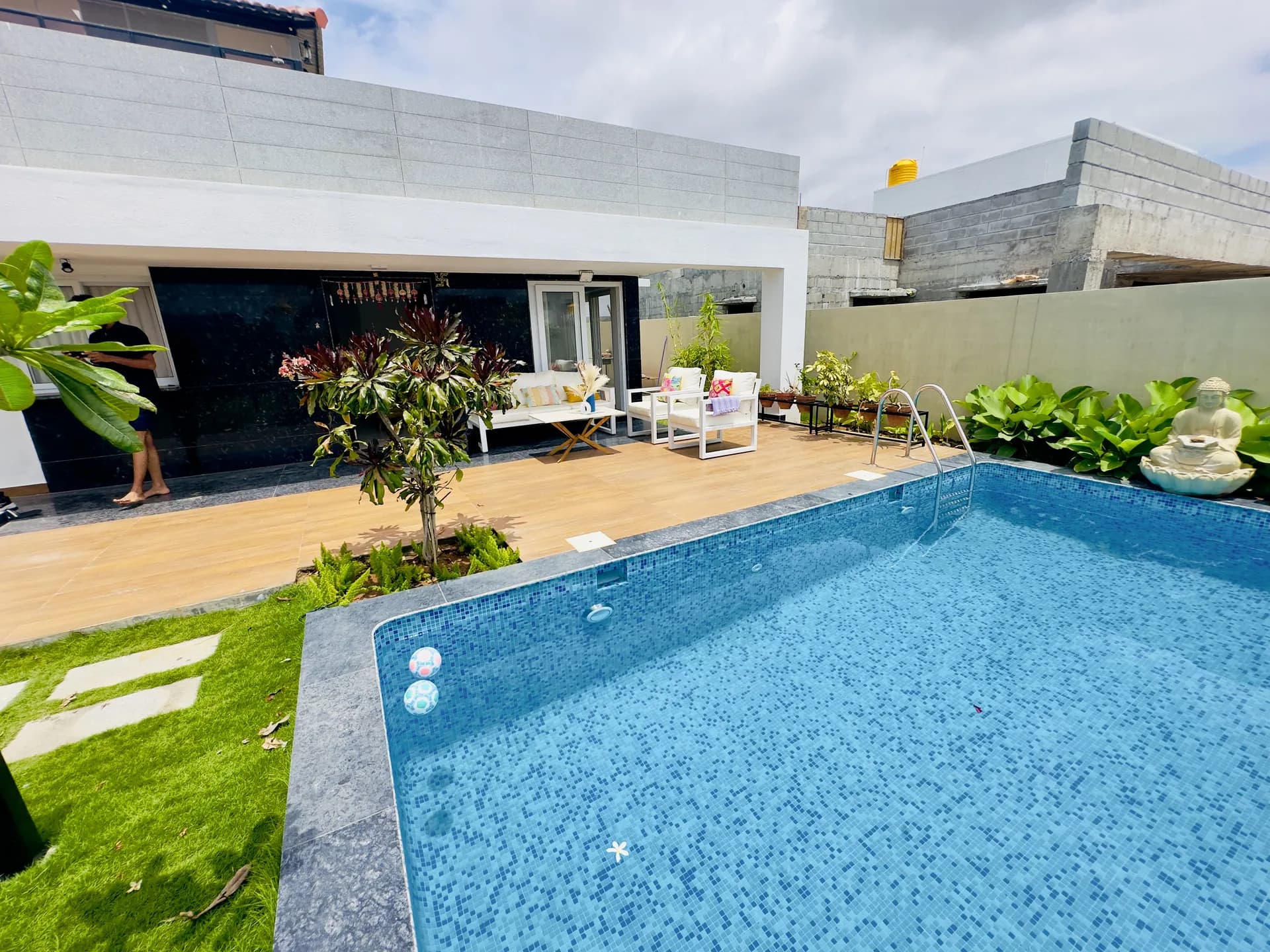 Outdoor swimming pool with blue mosaic tiles beside a timber deck, white lounge chairs and a Buddha statue.