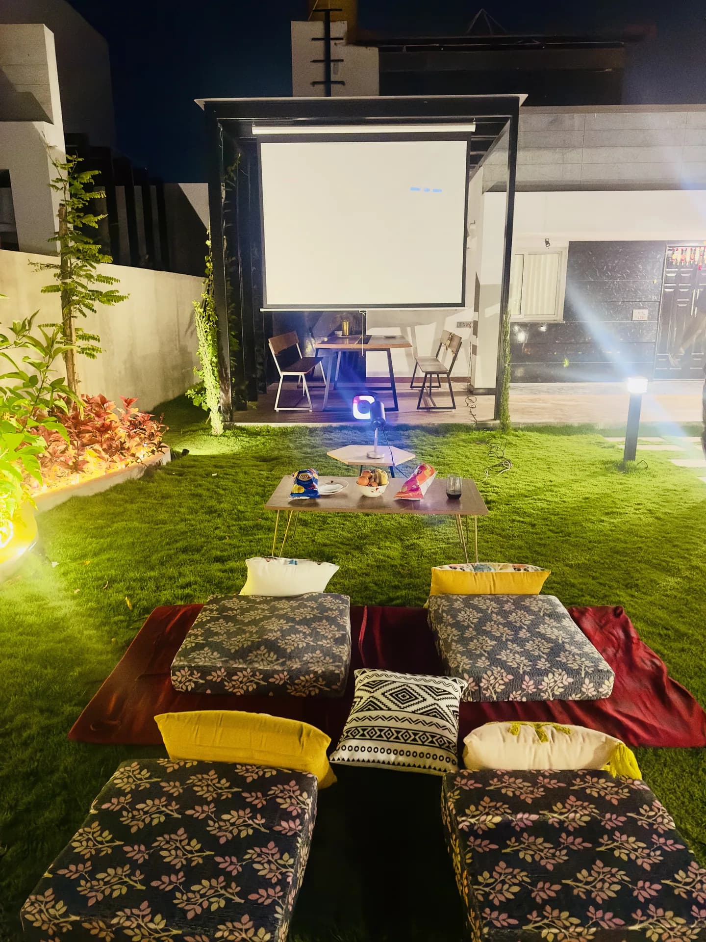 Outdoor cinema on lawn with projection screen, floor cushions, low wooden table and garden lighting at night.