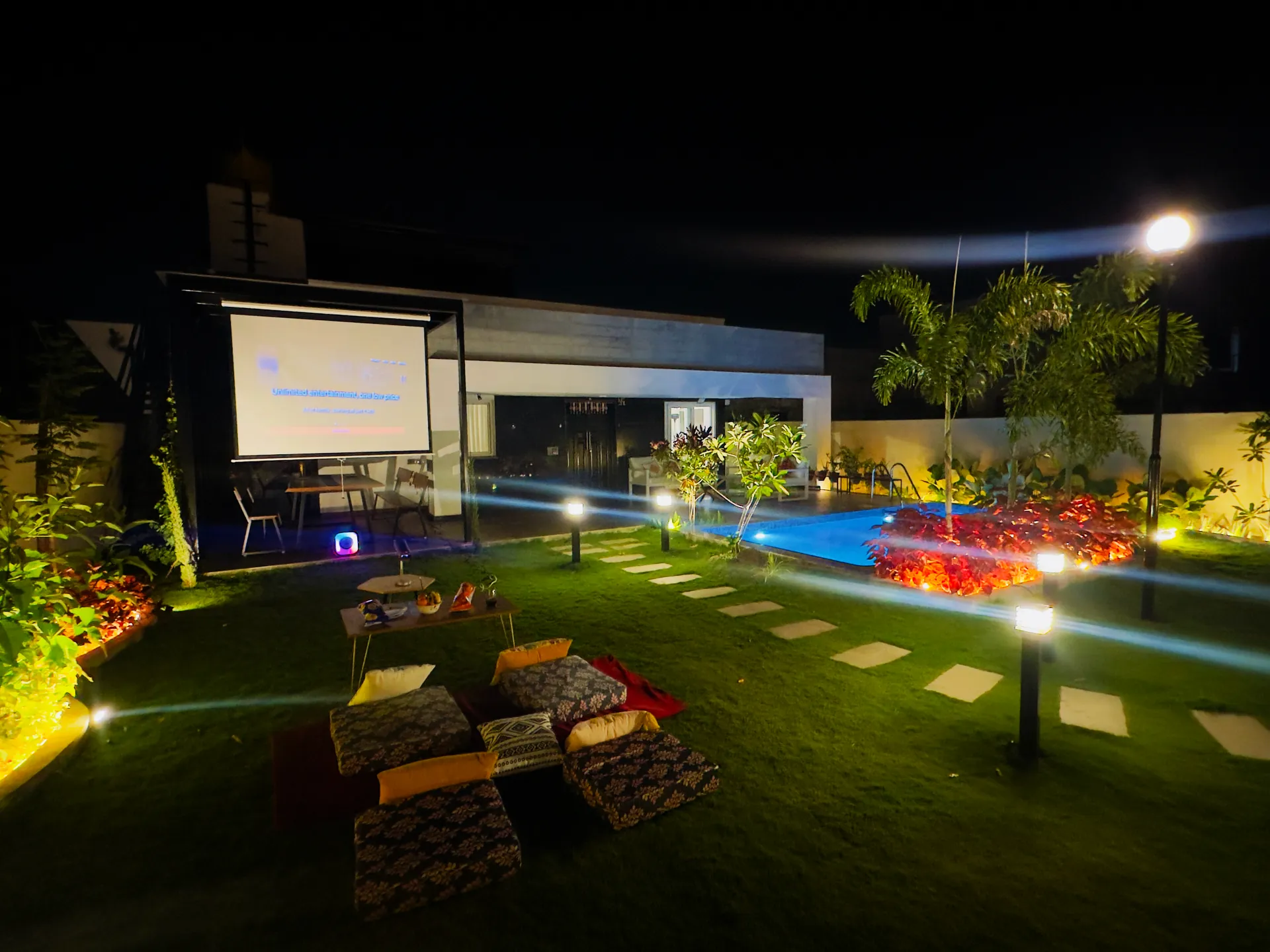 Night garden with a projector screen, floor cushions on lawn, and an illuminated swimming pool beside the villa.