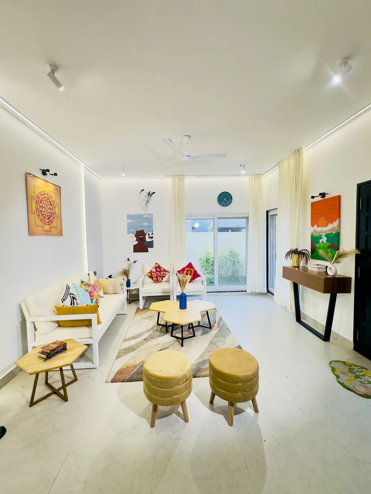Bright living room with white sofa, mustard stools, patterned rug, and glass doors opening to a garden.