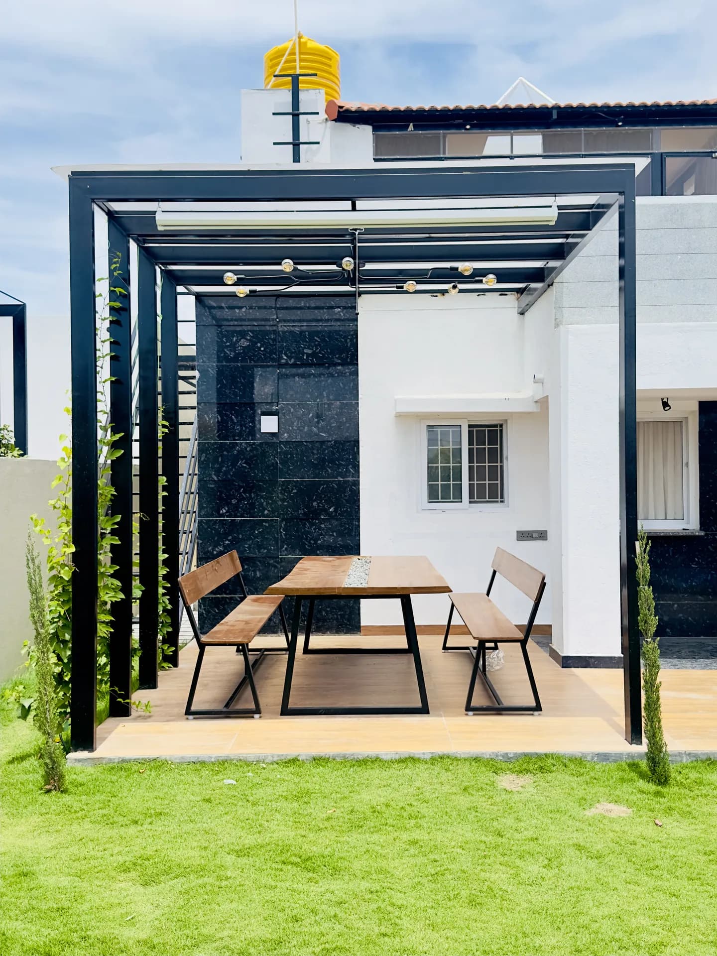 Covered outdoor patio with a wooden dining table, two benches, string lights and a lawn in front.