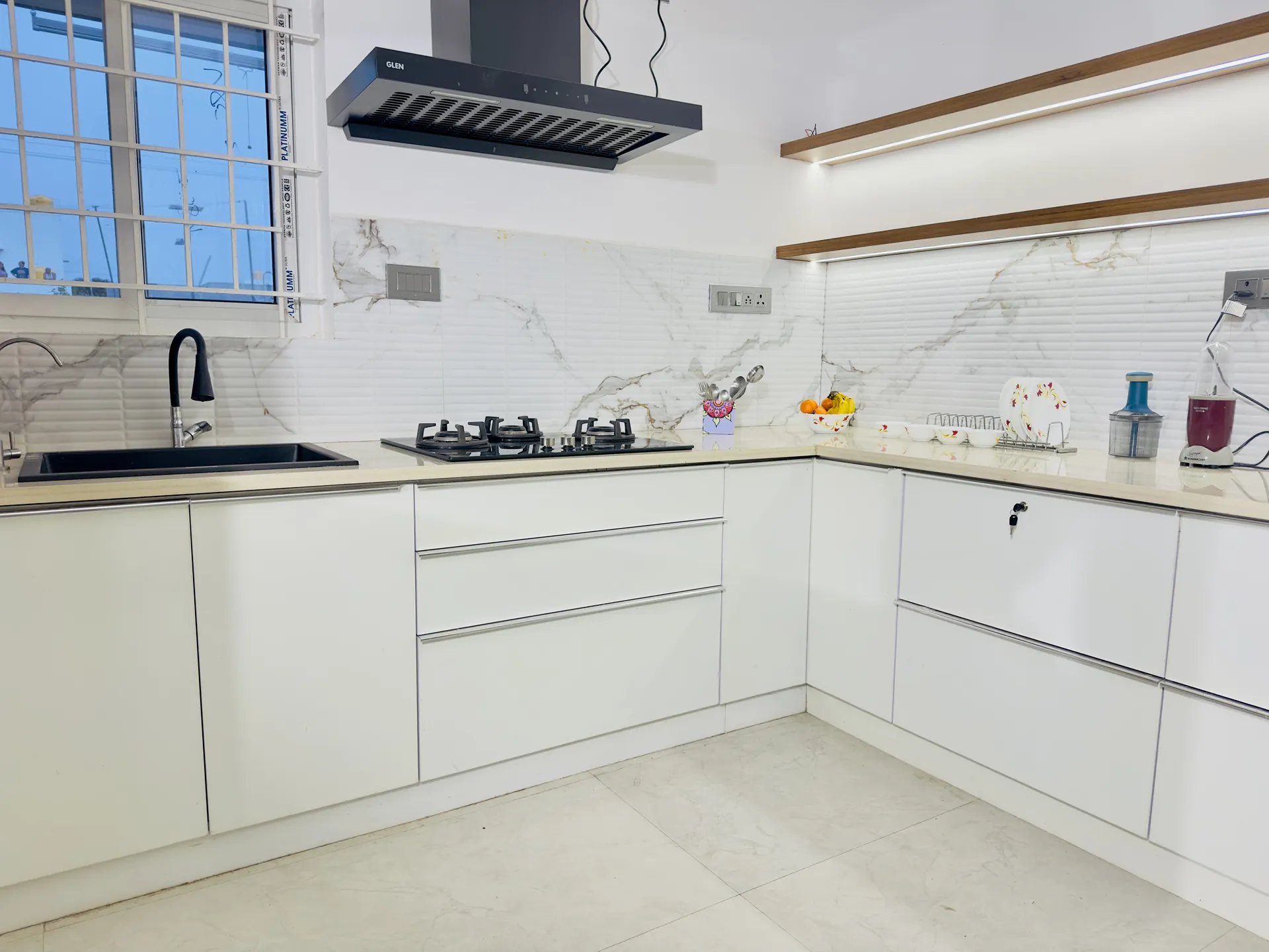 White kitchen with black gas hob, black sink, marble-look splashback and wooden shelf with under-lighting.