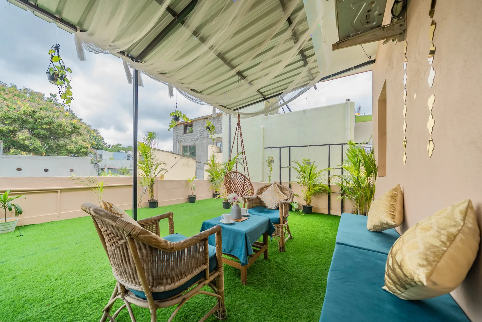 Covered terrace with artificial grass, wicker chairs, a blue-topped table, hanging swing chair and built-in cushioned bench.