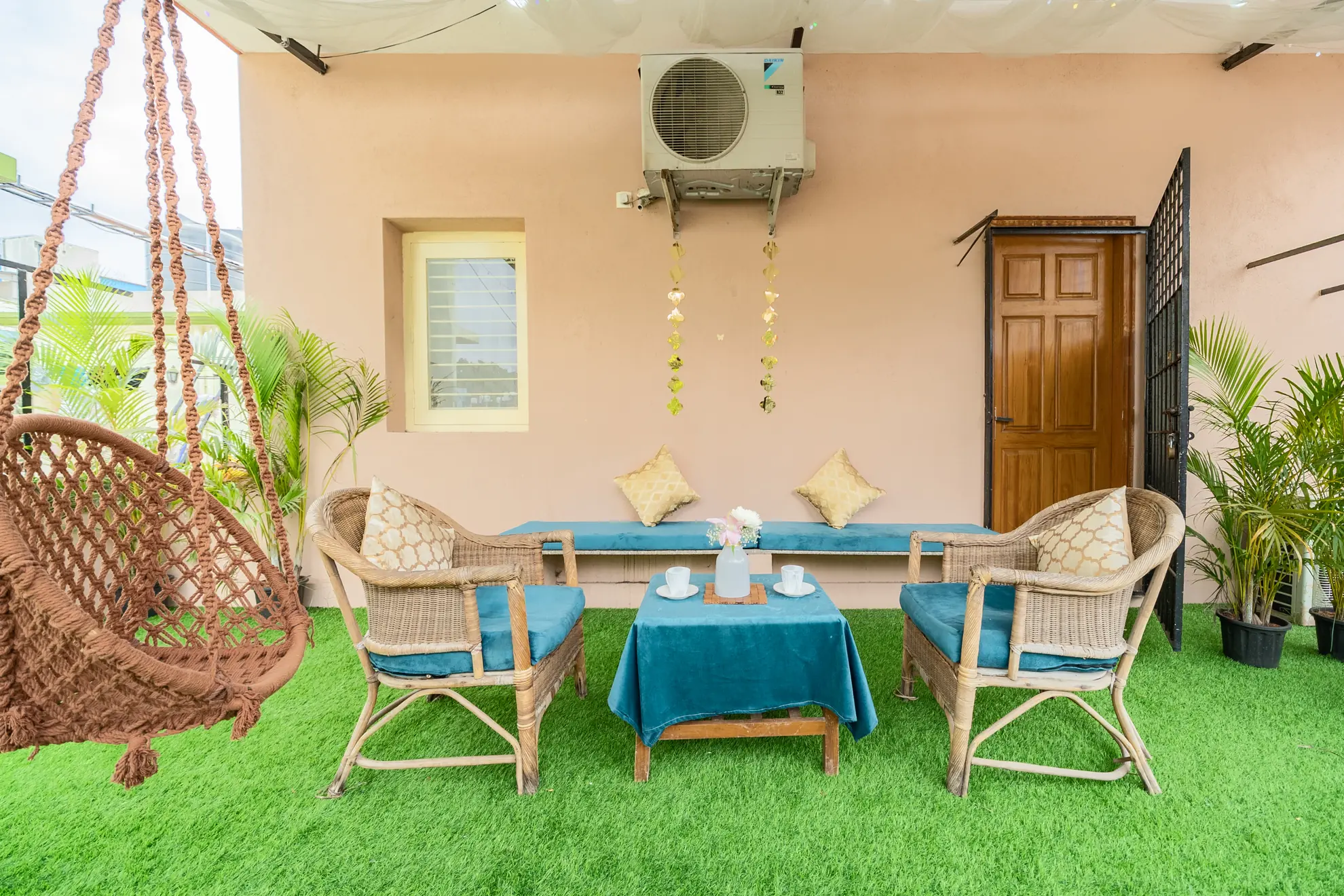 Outdoor terrace with artificial grass, wicker armchairs with teal cushions, a teal-covered coffee table and a hanging rope swing chair.