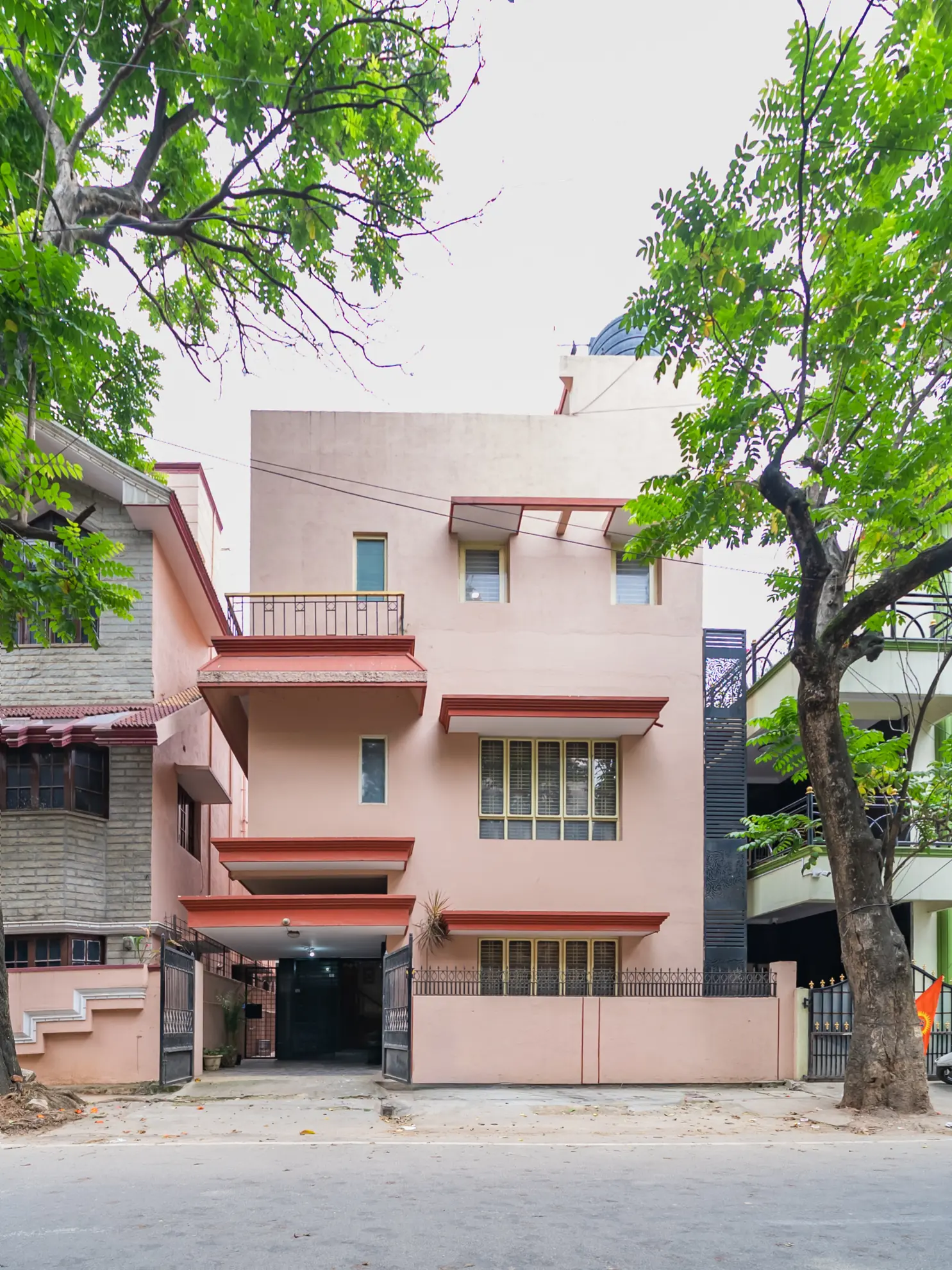 Pink three-storey villa facade behind a low wall, with a gated driveway entrance and carport at street level.