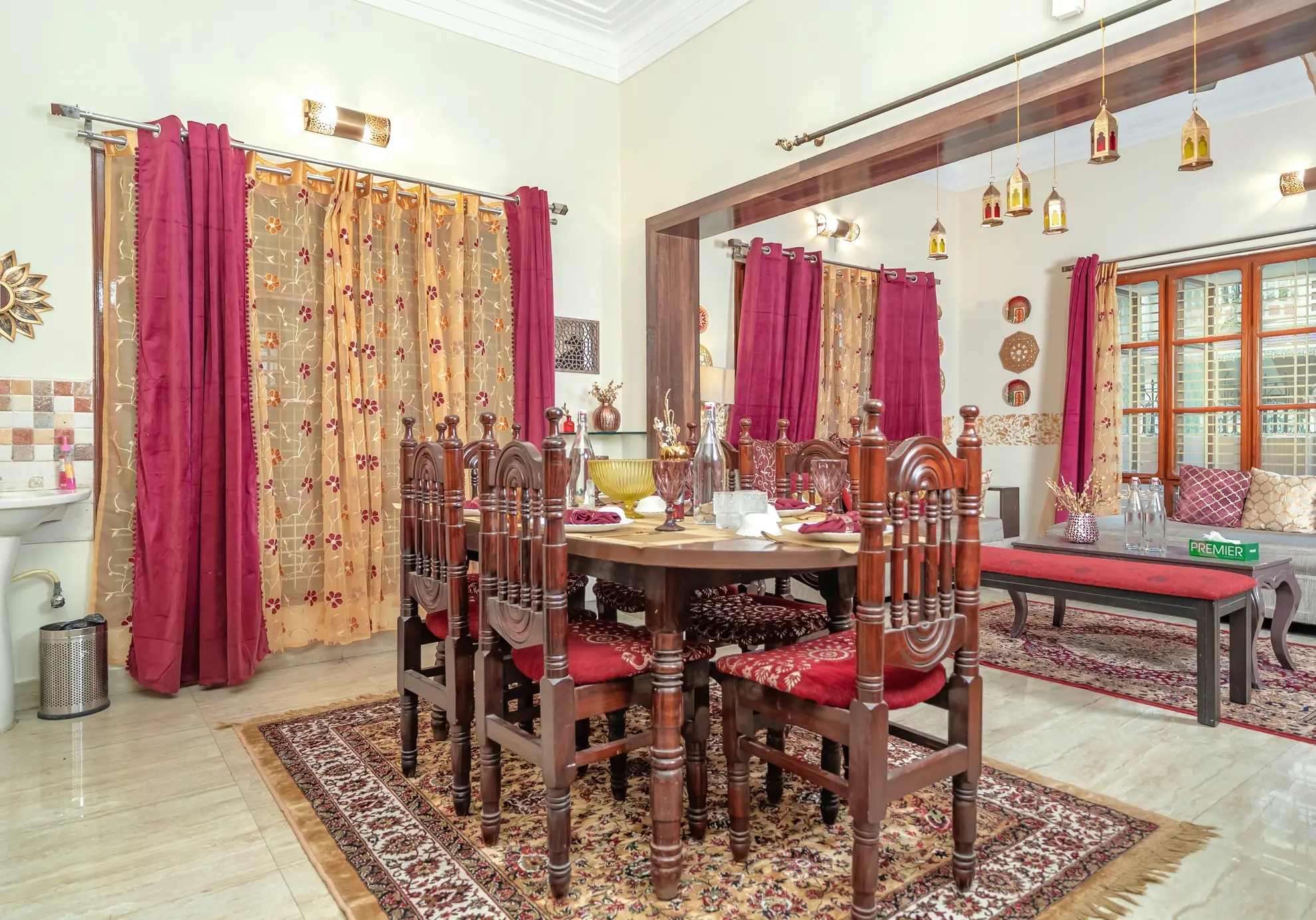 Dining area with a round wooden table and carved chairs, red and gold curtains, and a low daybed beside tall windows.