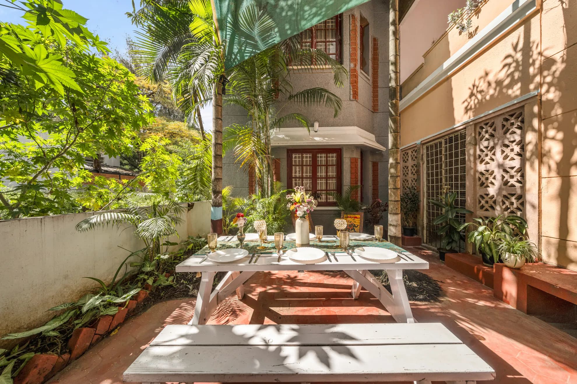 Outdoor courtyard patio with a set dining table under a shade sail, lush plants and a white brick-trim building behind.