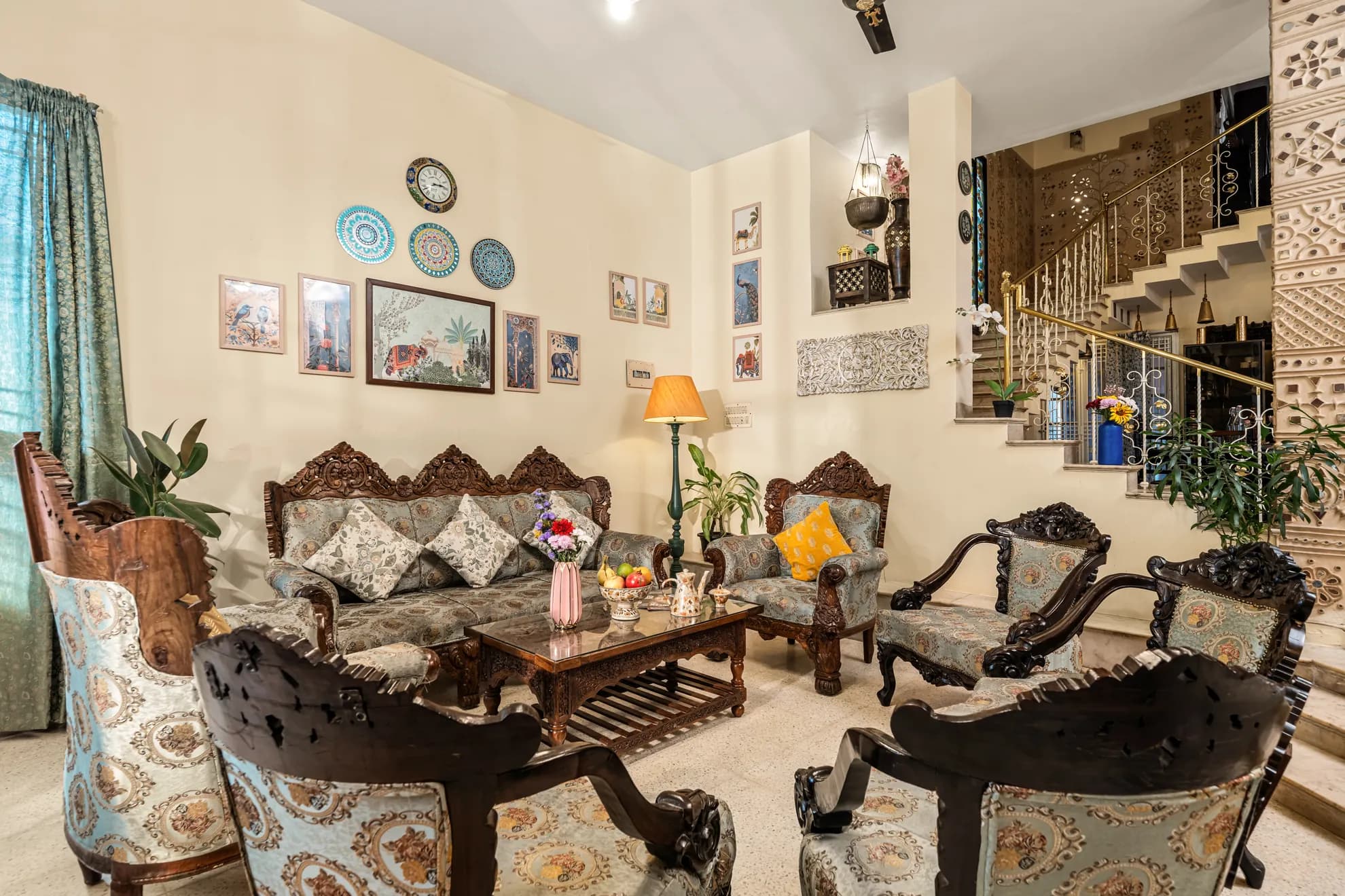 Living room with carved wooden sofa and armchairs, a wall of decorative plates and framed prints, and a staircase with ornate railings.