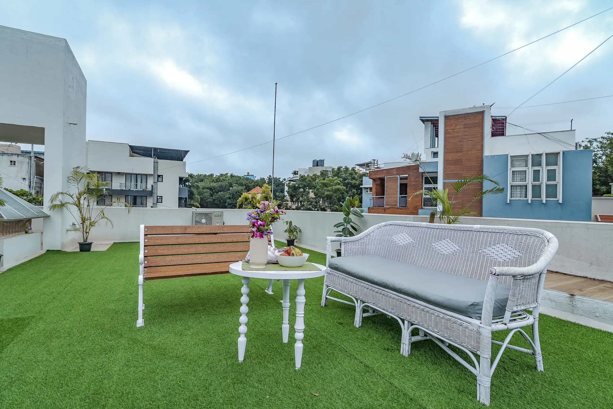 Rooftop terrace with artificial turf, white wicker sofa, round side table with flowers and fruit, and a wooden bench.