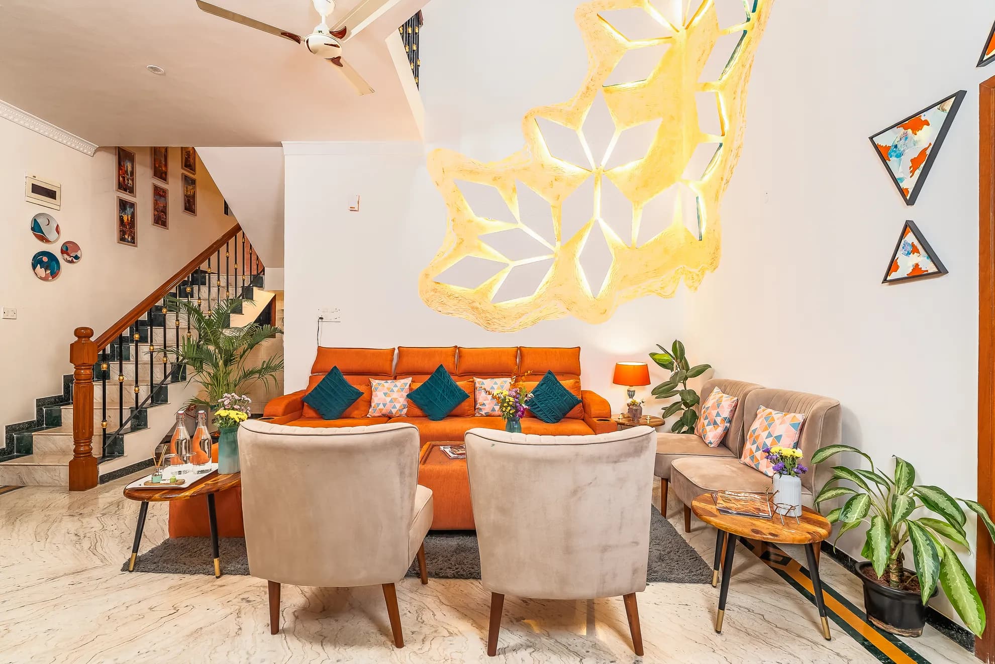 Living room with an orange sofa, two cream armchairs, a lit sculptural wall lamp and a staircase.