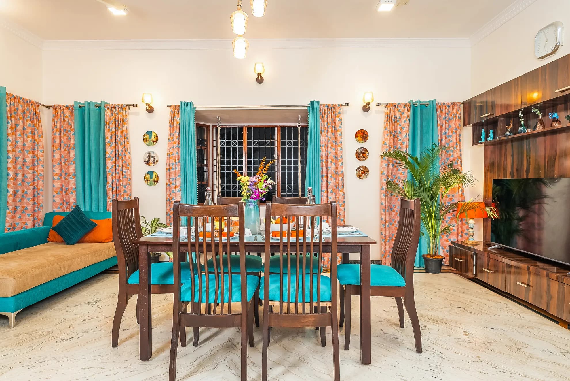 Bright dining area with a wooden table for six, teal chairs, and a sofa with orange cushions beside it.