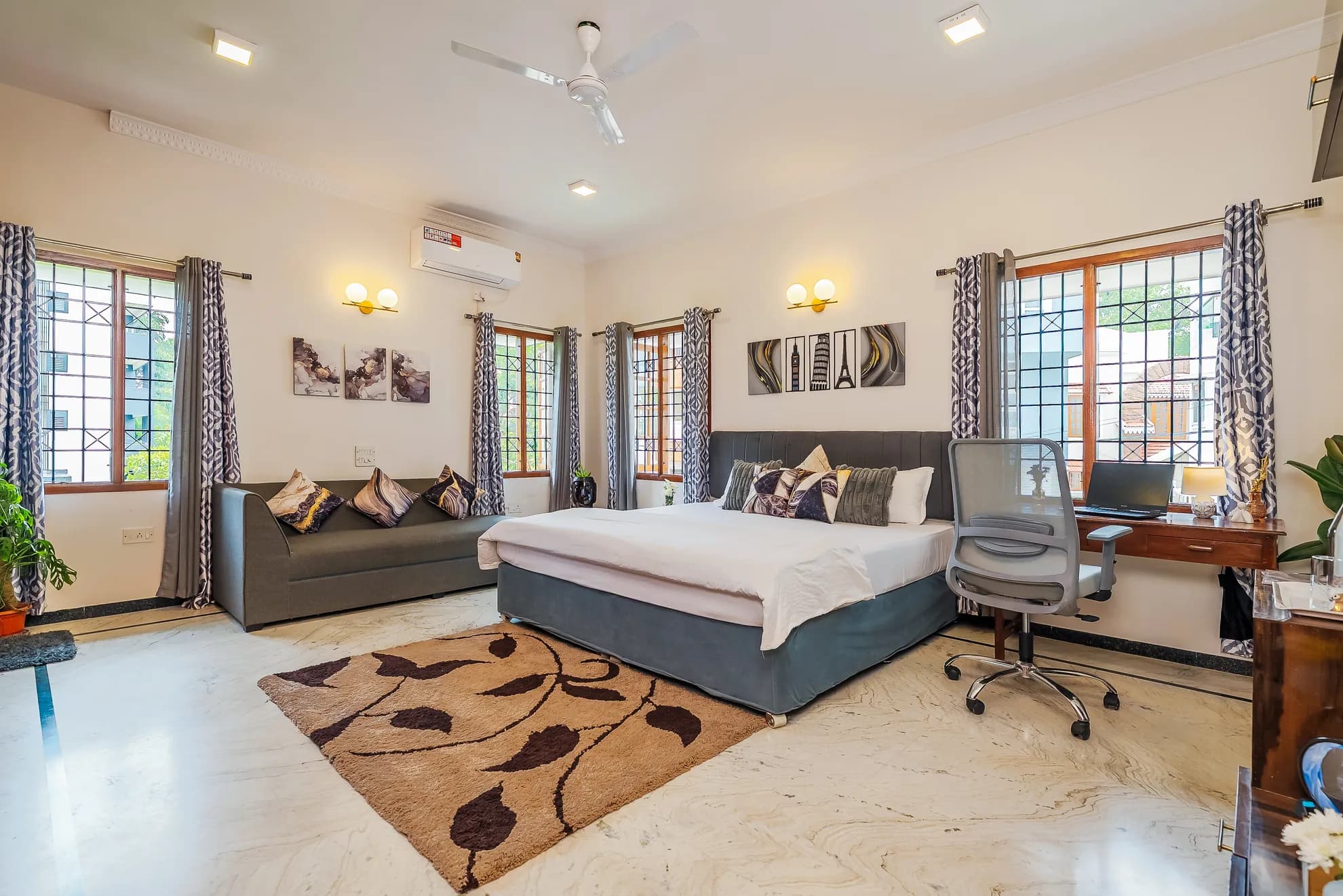 Large bedroom with a king bed, grey sofa, wall-mounted AC unit, and a wooden desk with a chair by the window.