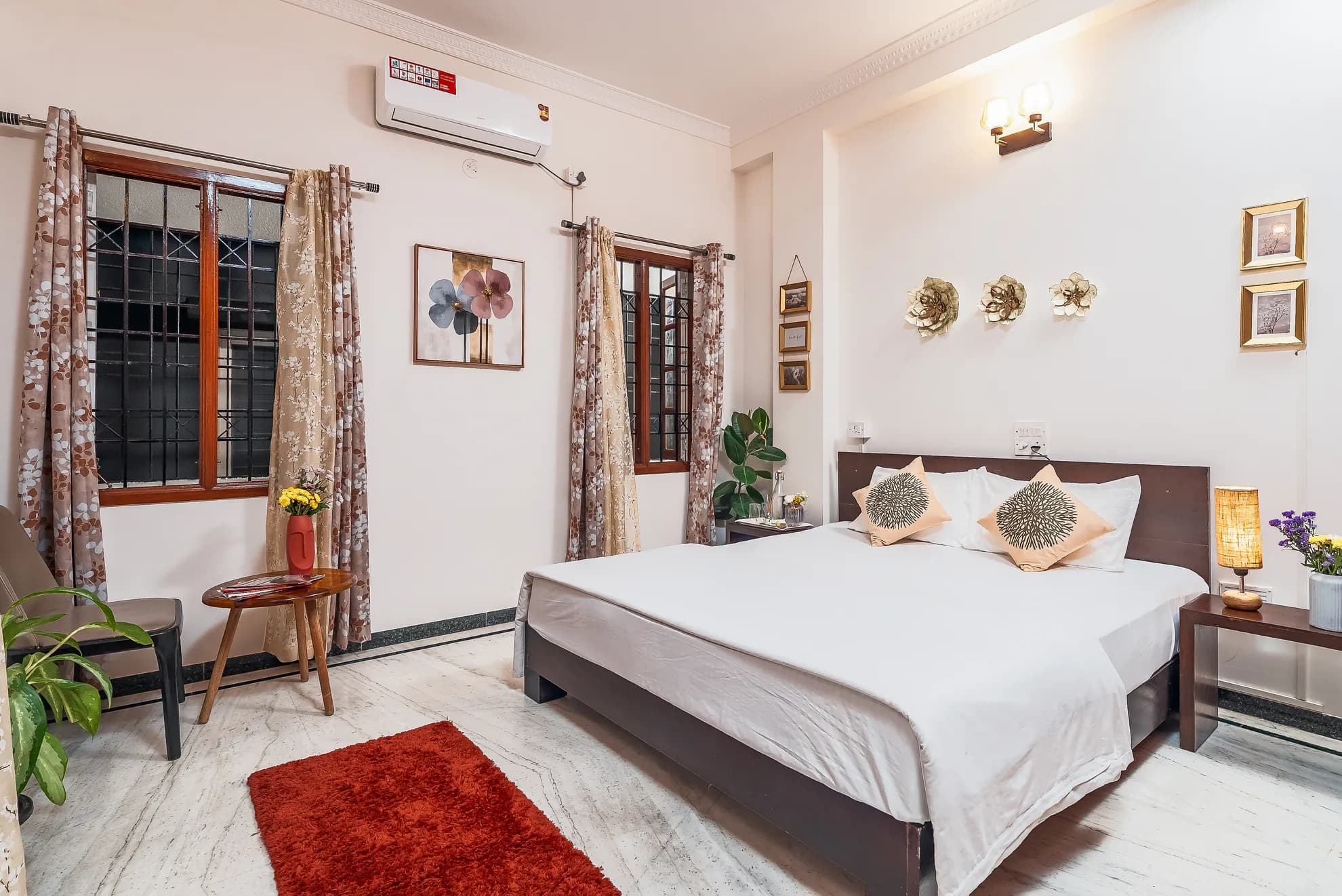 Bedroom with a made-up double bed, wall air conditioner, two barred windows with patterned curtains, and a red rug.