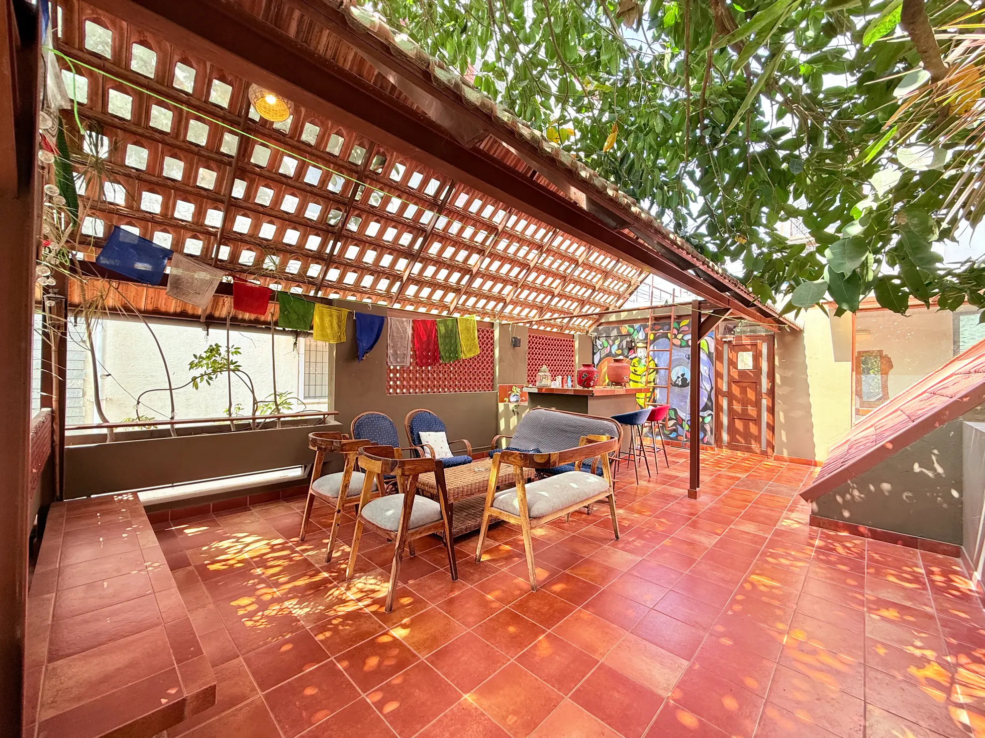 Covered terrace with red terracotta tiles, wooden chairs, a low sofa and a bar counter under a lattice roof.