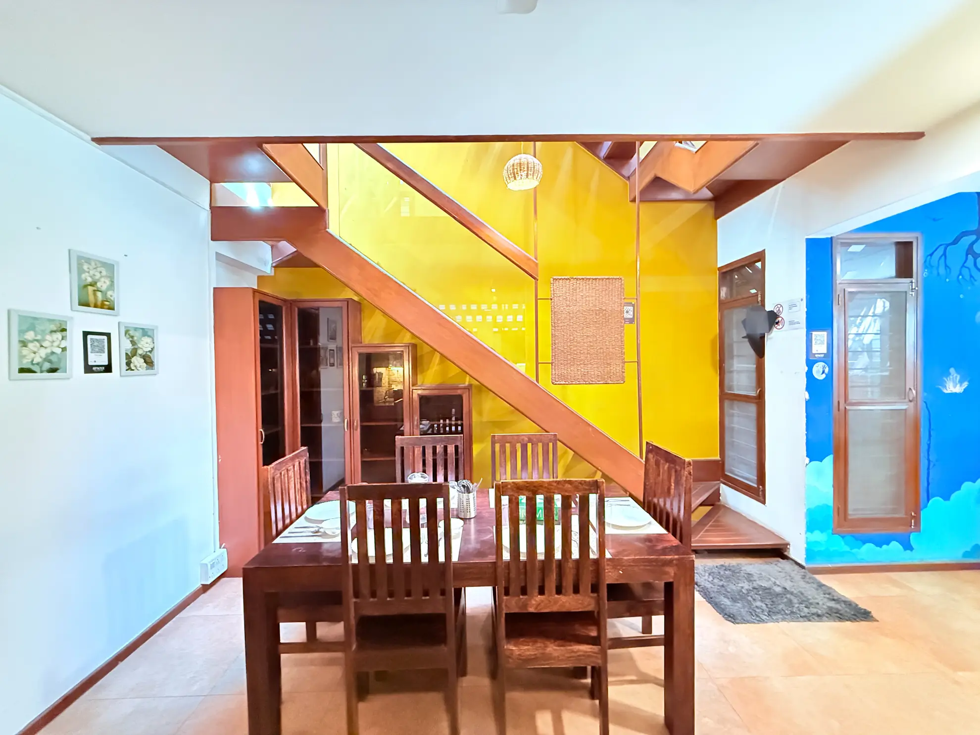 Dining area with a wooden table and chairs, a yellow staircase wall and a blue mural beside the door.