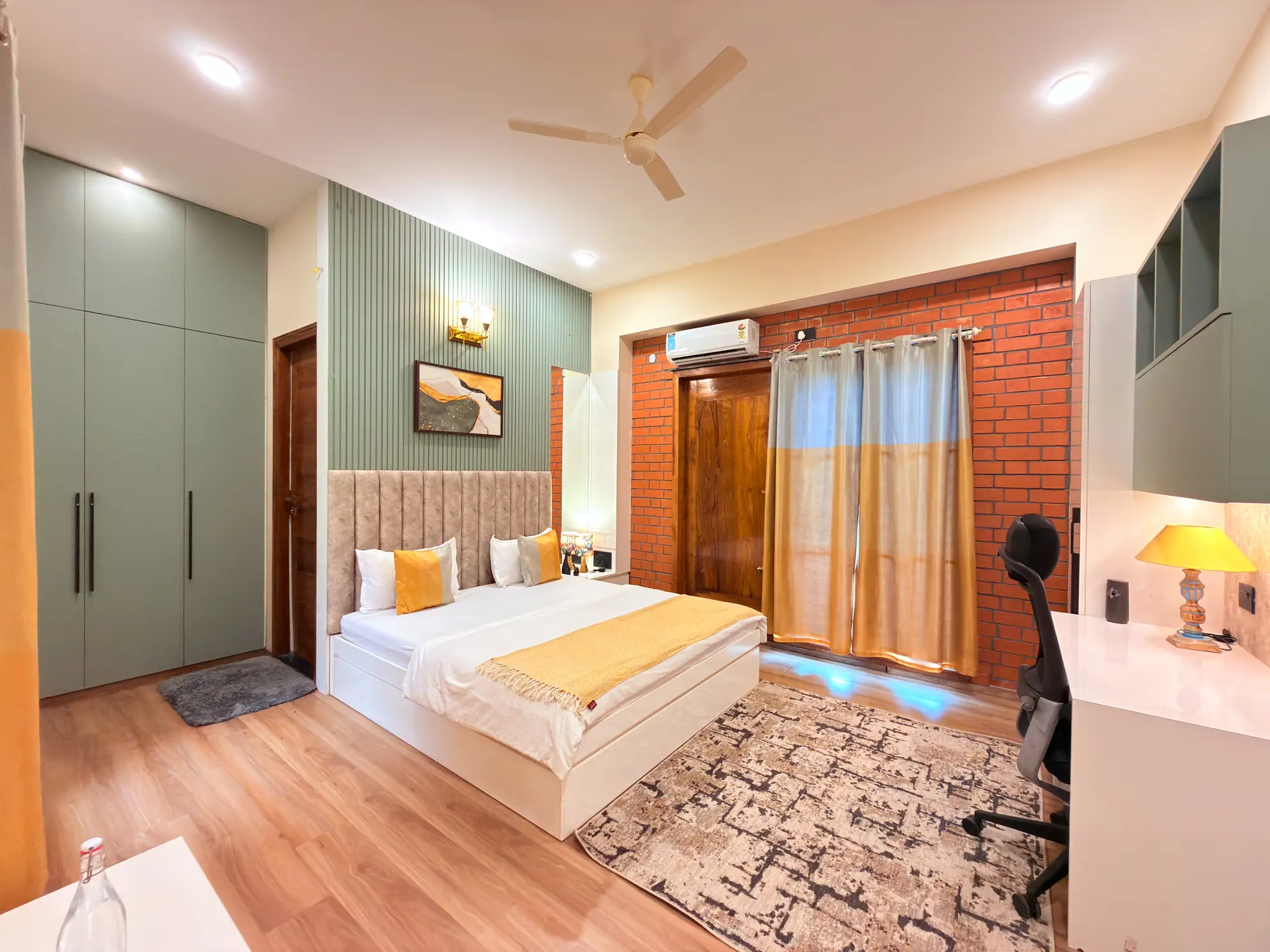 Bedroom with a made bed, green wardrobe, exposed brick wall, ceiling fan and a wall-mounted air conditioner.