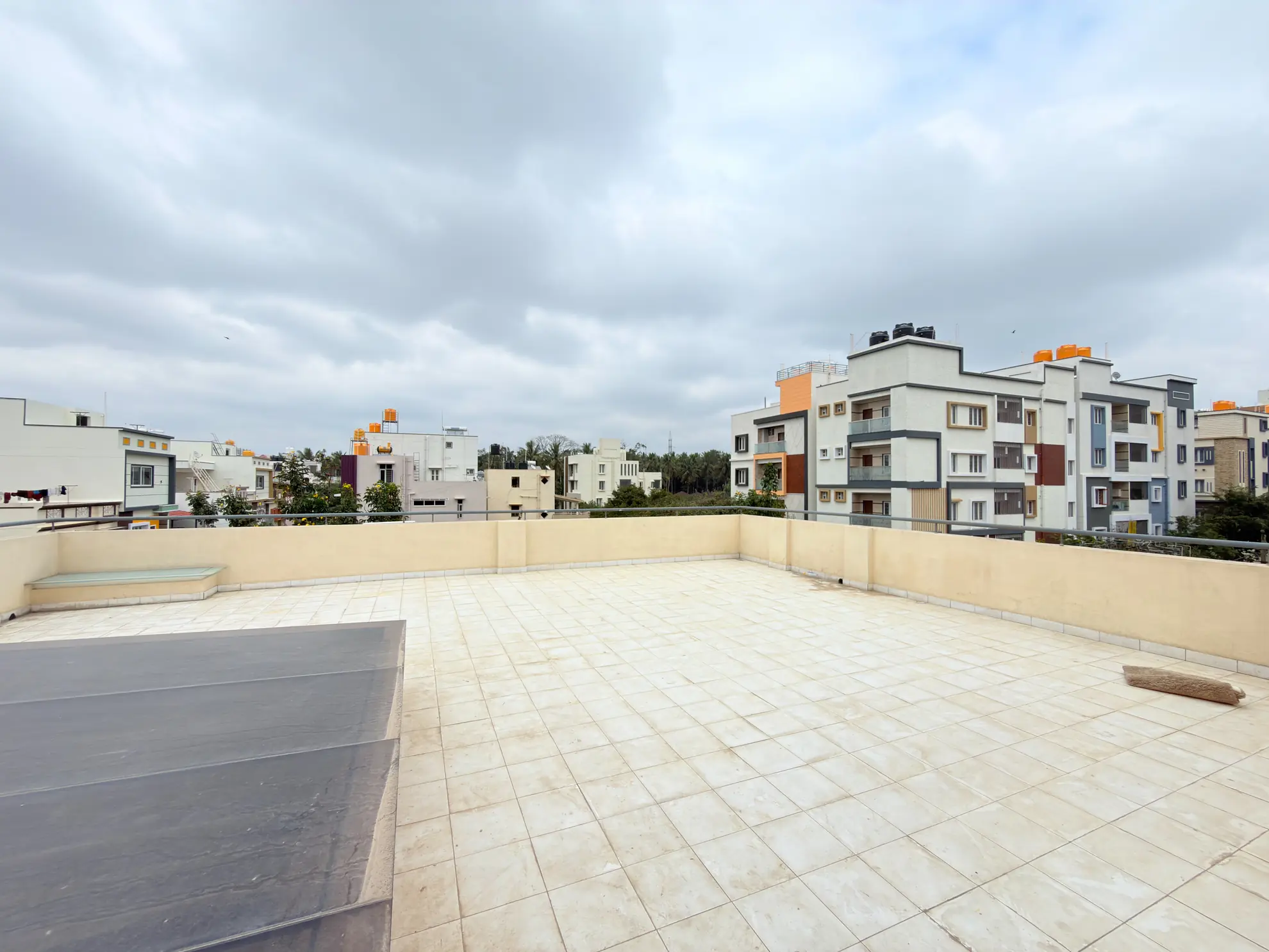 Open rooftop terrace with cream tiled floor, low parapet walls, and neighbouring apartment buildings beyond under an overcast sky.