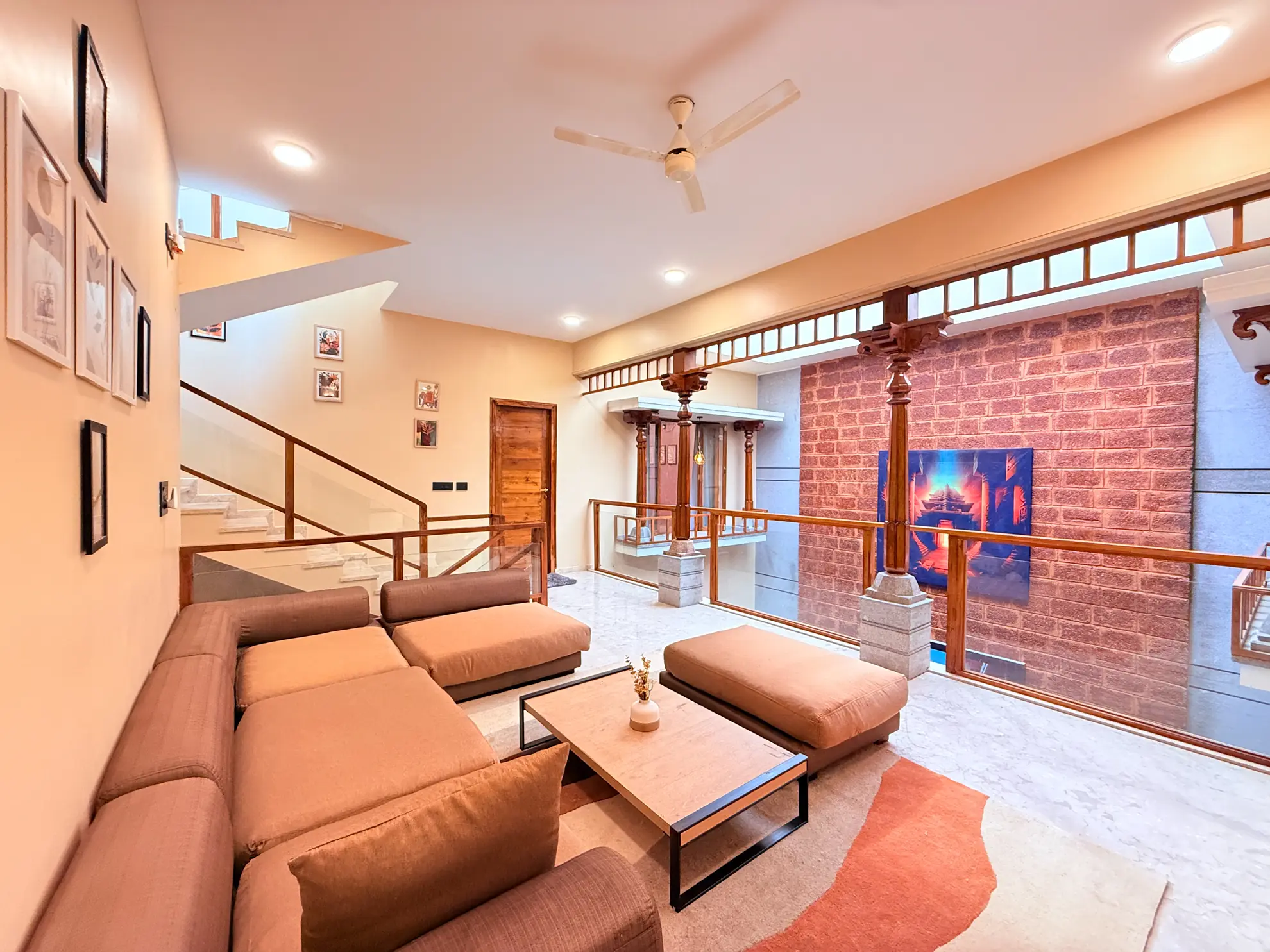 Open-plan lounge with tan sectional sofa, wood-railed staircase, and exposed brick wall beside a ceiling fan.