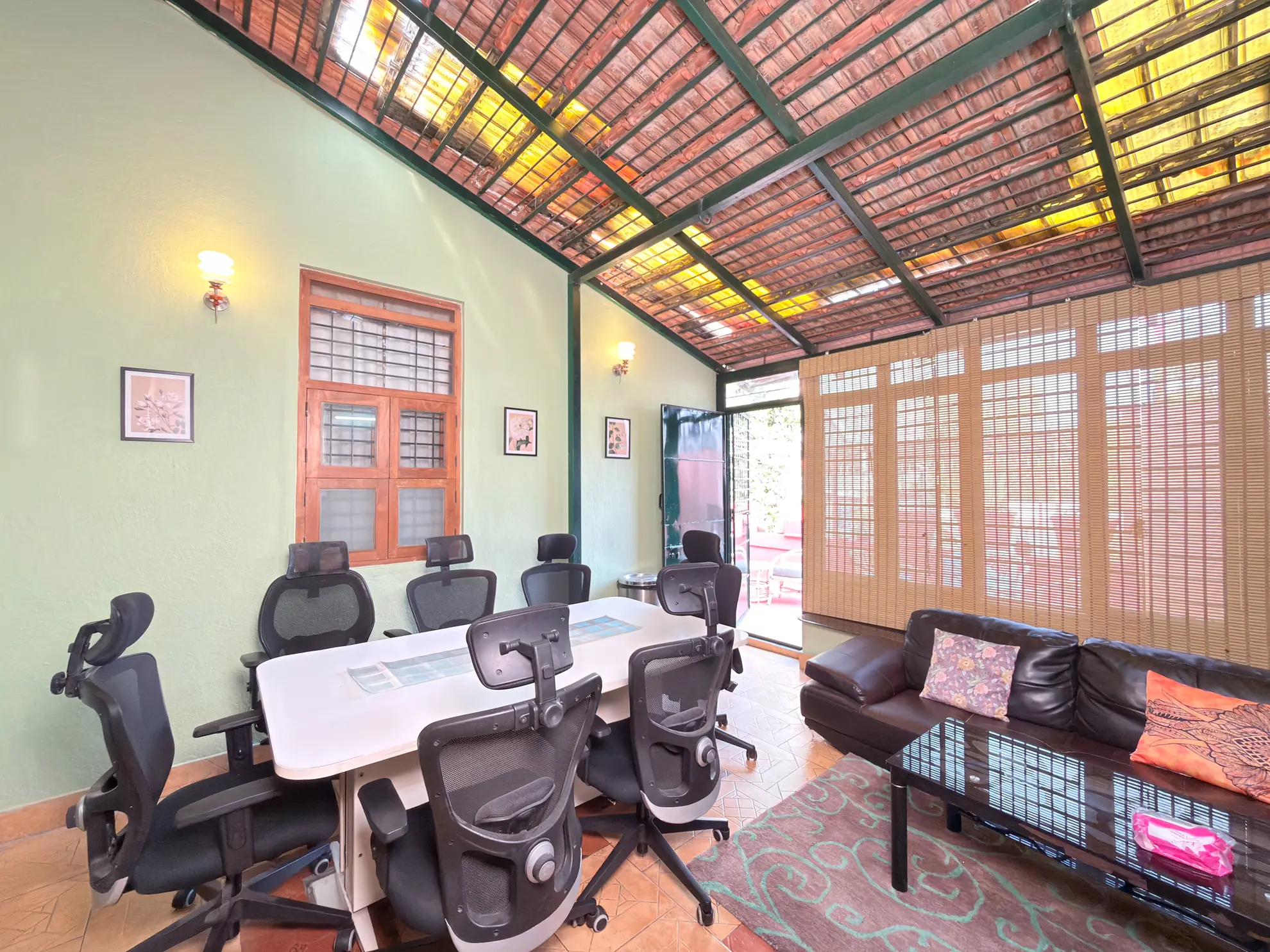 Covered workspace with a white conference table, black mesh office chairs, and a leather sofa beside a bamboo blind.
