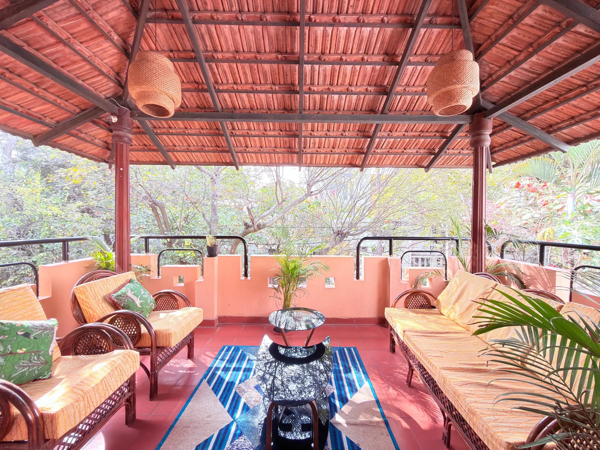 Covered verandah with two wicker sofas, rattan armchairs, a glass side table, and a blue patterned rug under a terracotta roof.