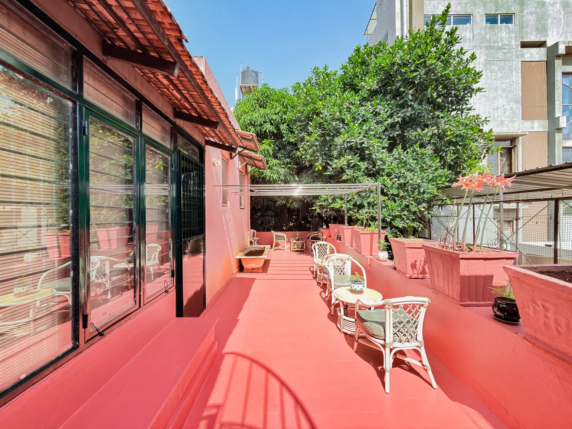 Red-tiled terrace with white wicker chairs, a small round table, potted plants and a large shade tree beside it.